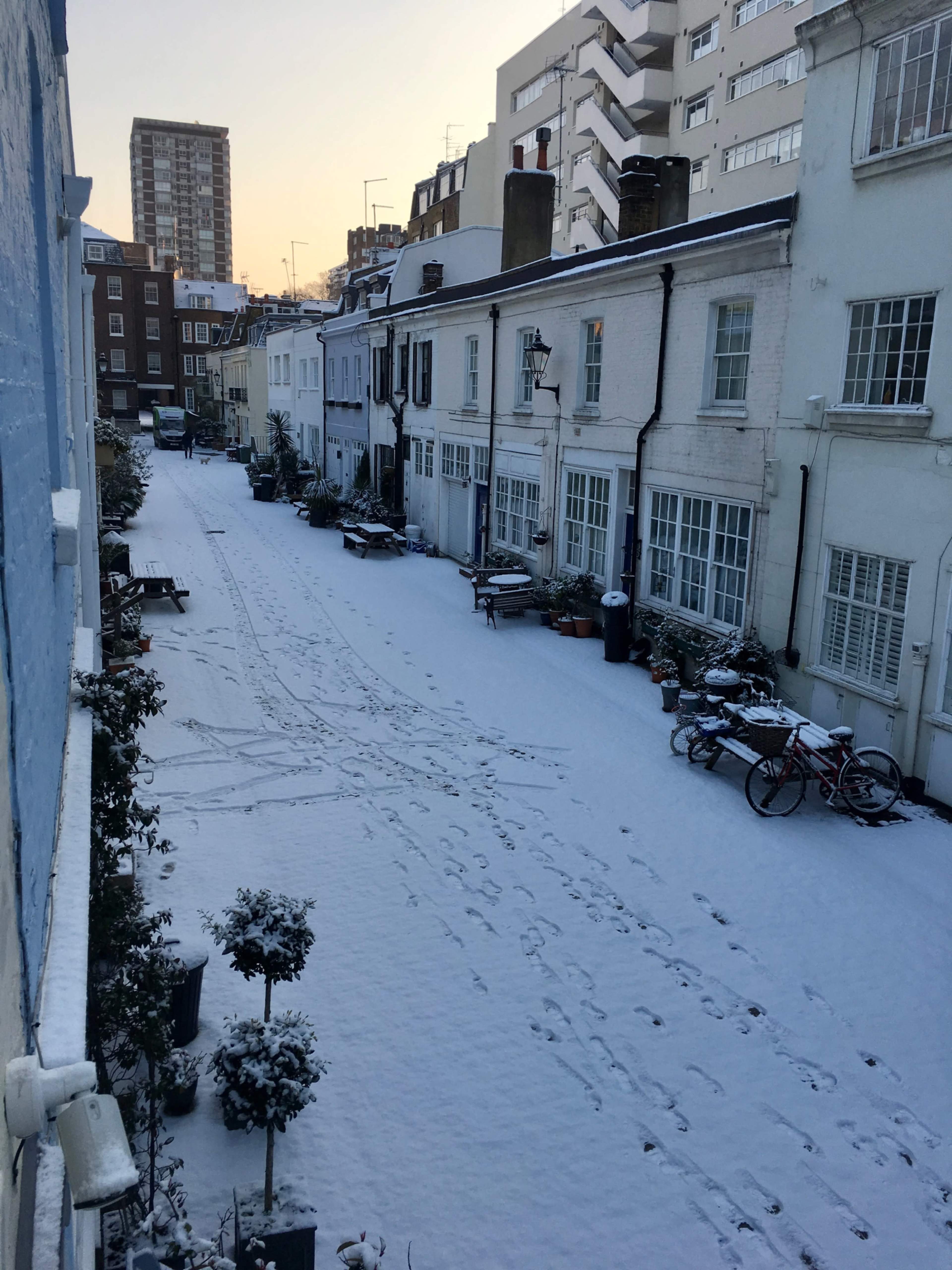 A snow-covered narrow street lined with modest houses and scattered bicycles in a residential area.