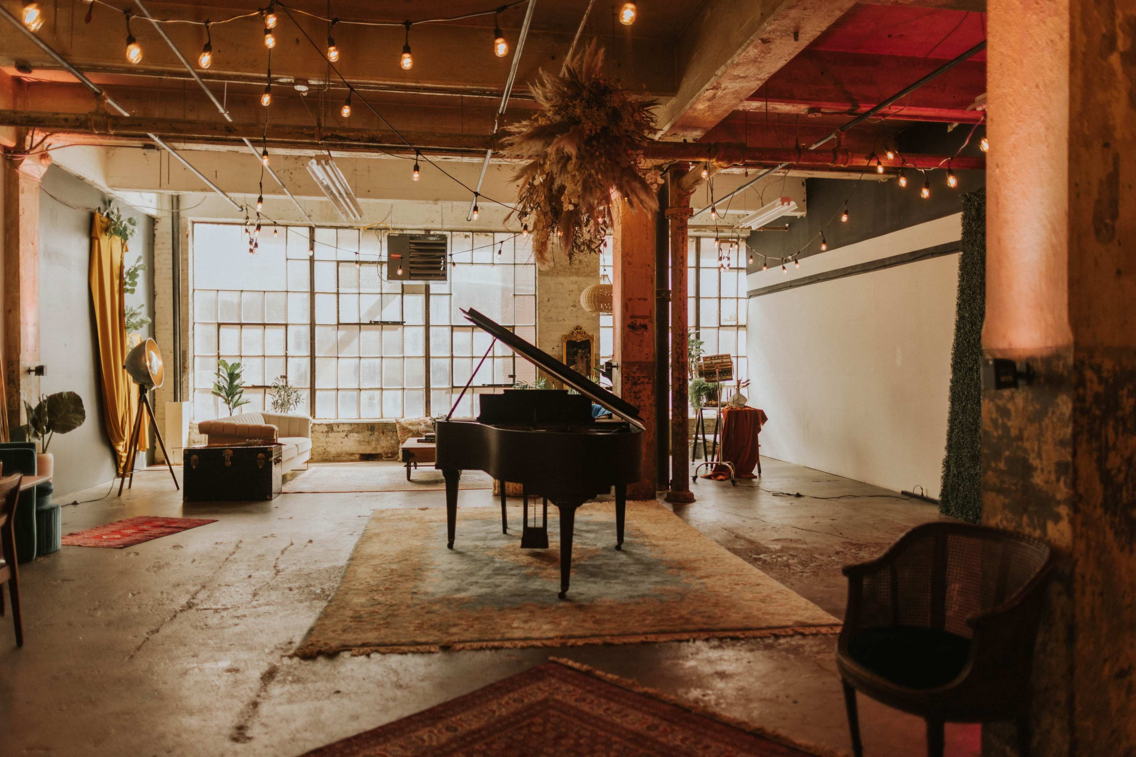 A grand piano sits on a patterned rug in a spacious room with string lights and large windows.