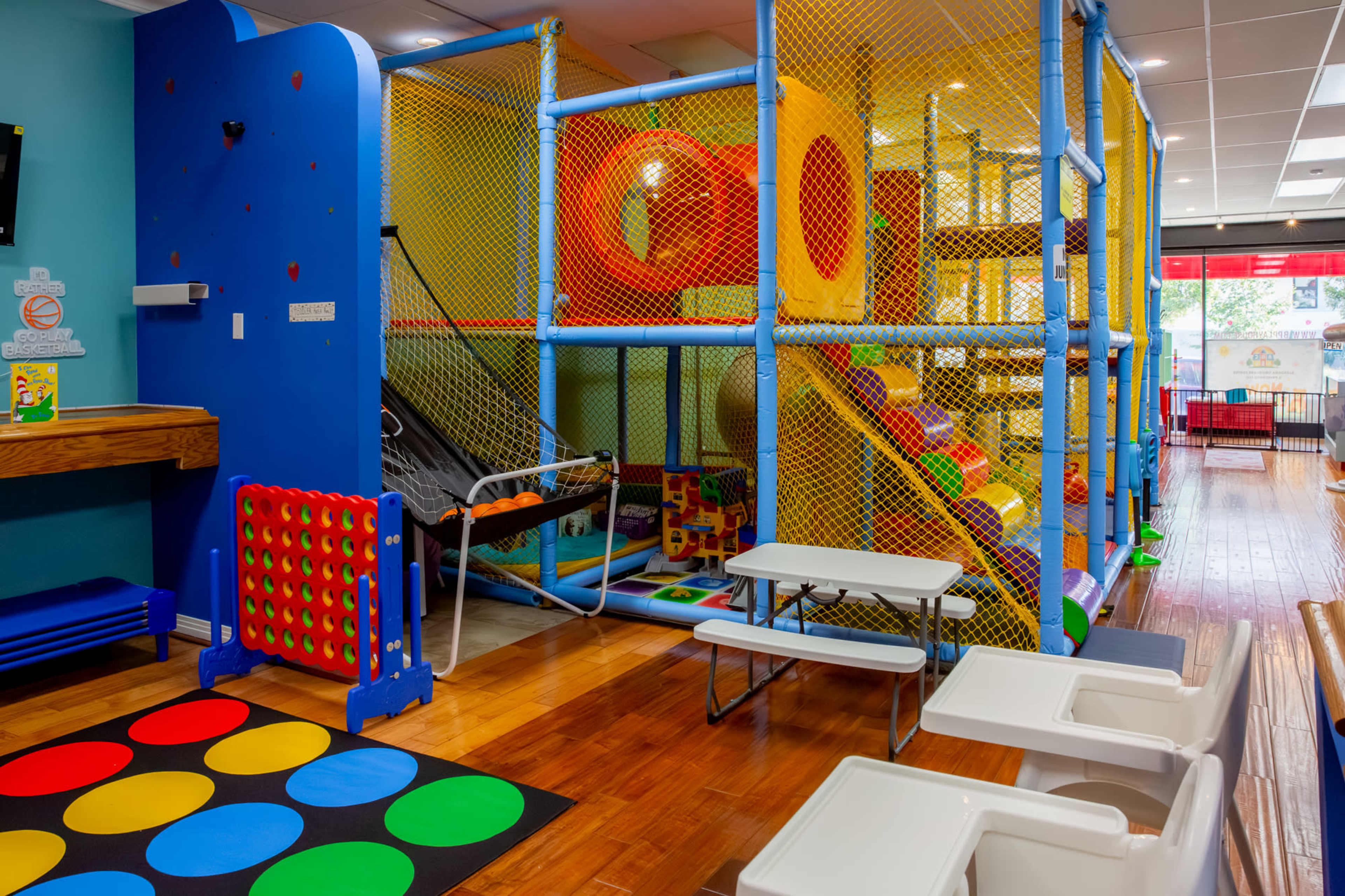 The image shows a colorful indoor play area with climbing structures, slides, and soft play equipment, surrounded by safety netting.