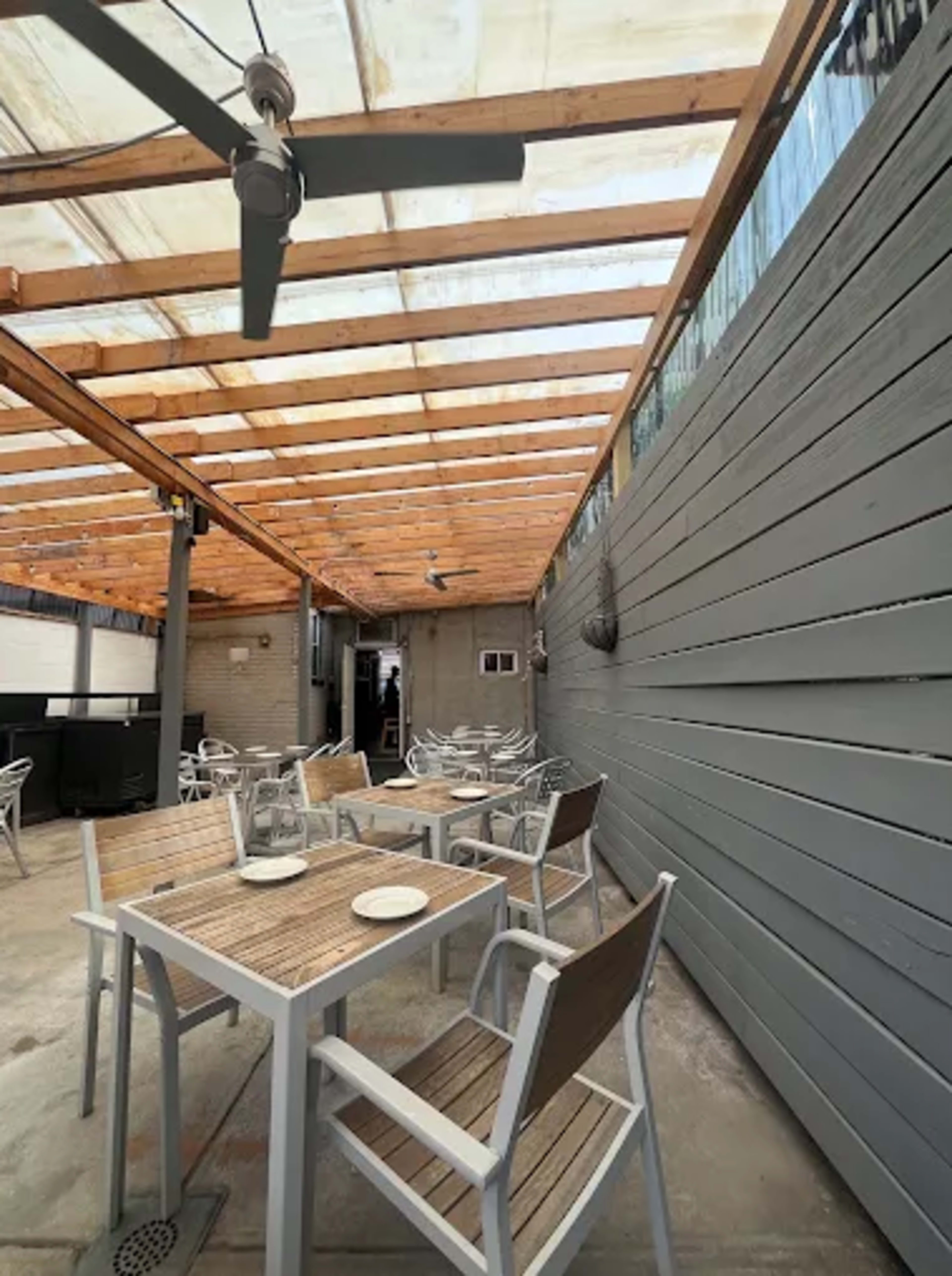 The image shows an outdoor dining area with wooden beams overhead, empty tables and chairs arranged on a paved floor, and a wall covered in gray panels.