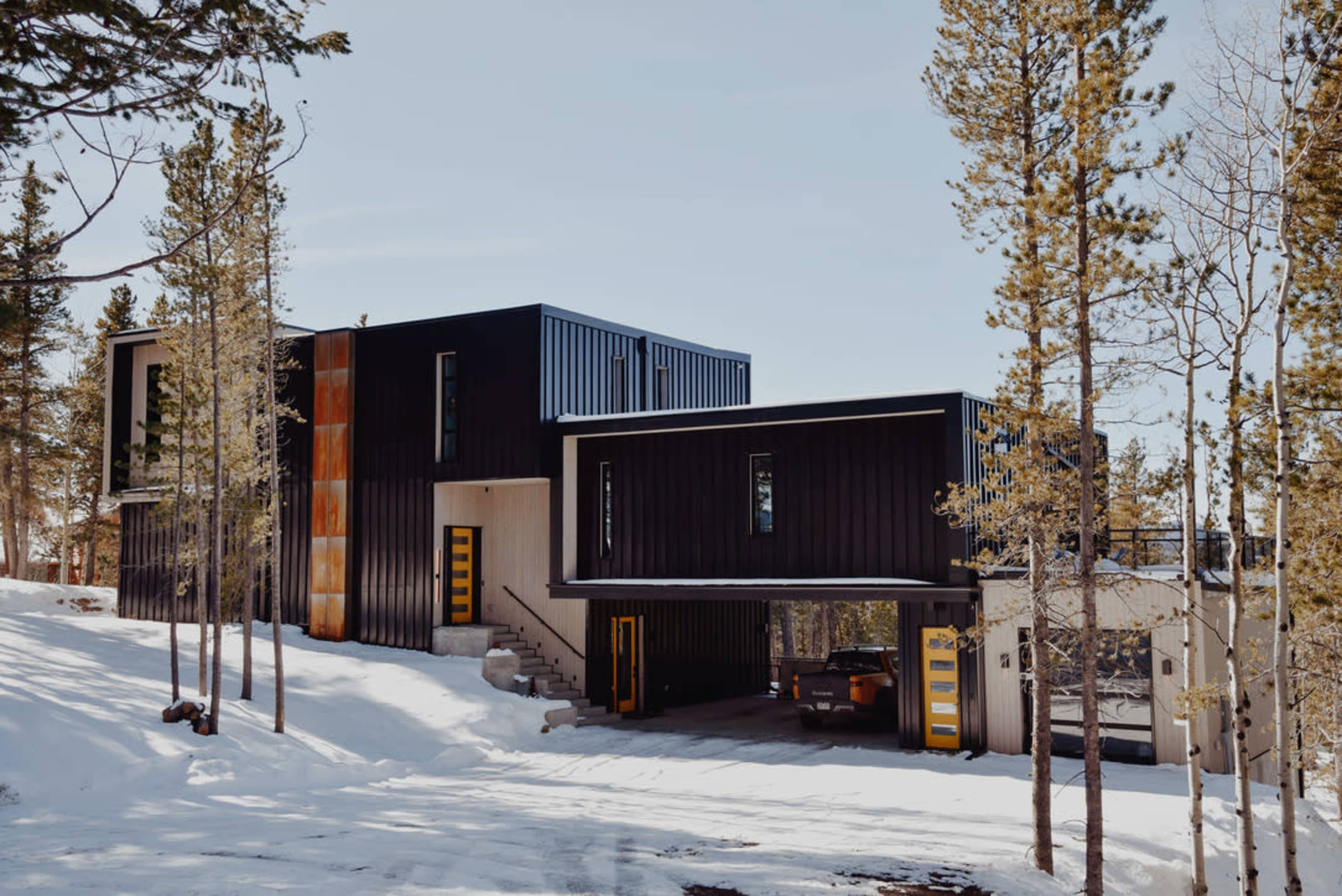 A modern two-story house with a mix of dark and light exterior materials is situated among snow-covered trees.