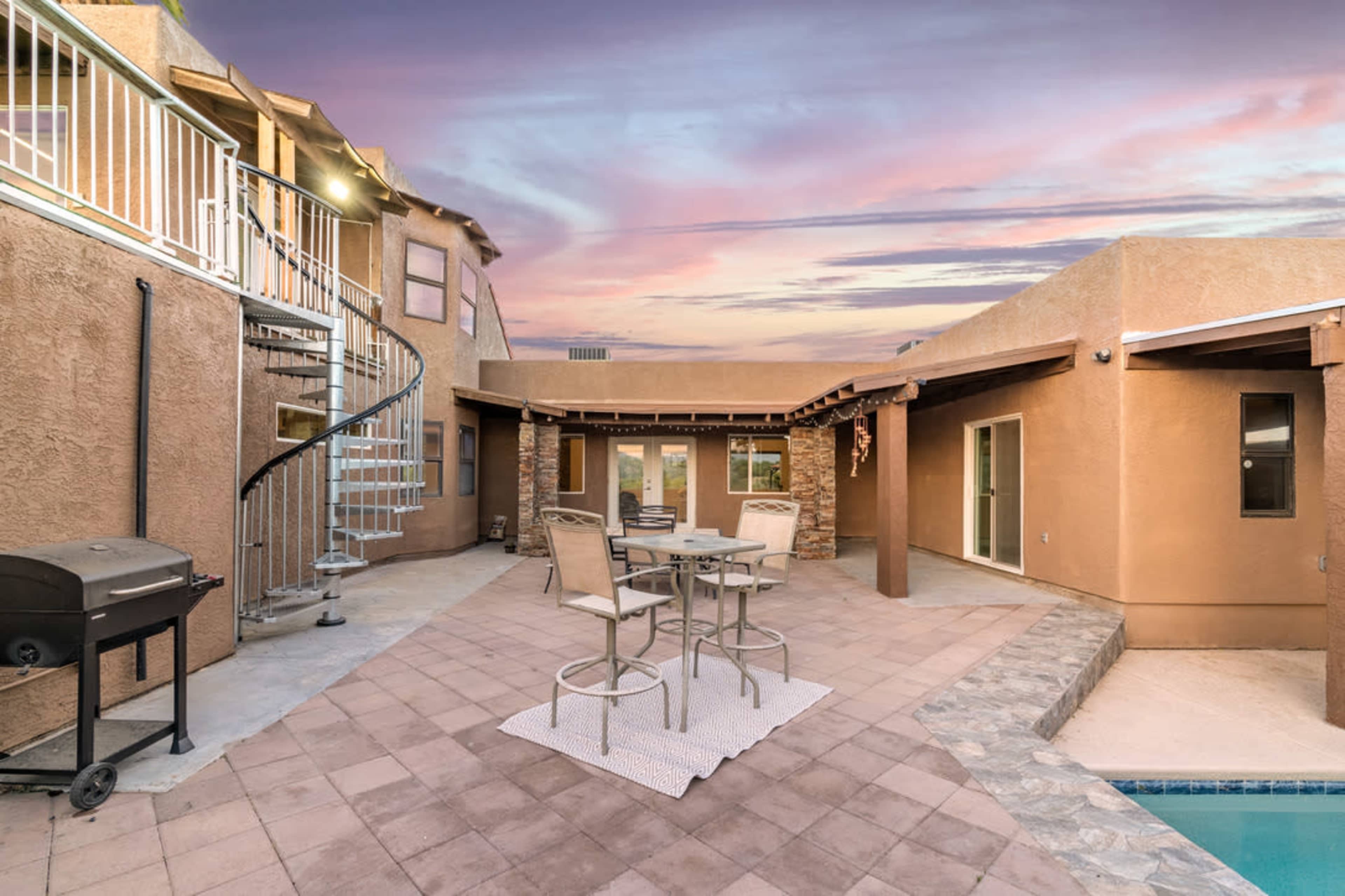 A courtyard with a spiral staircase, outdoor seating area, and a small pool, set against a colorful sunset sky.