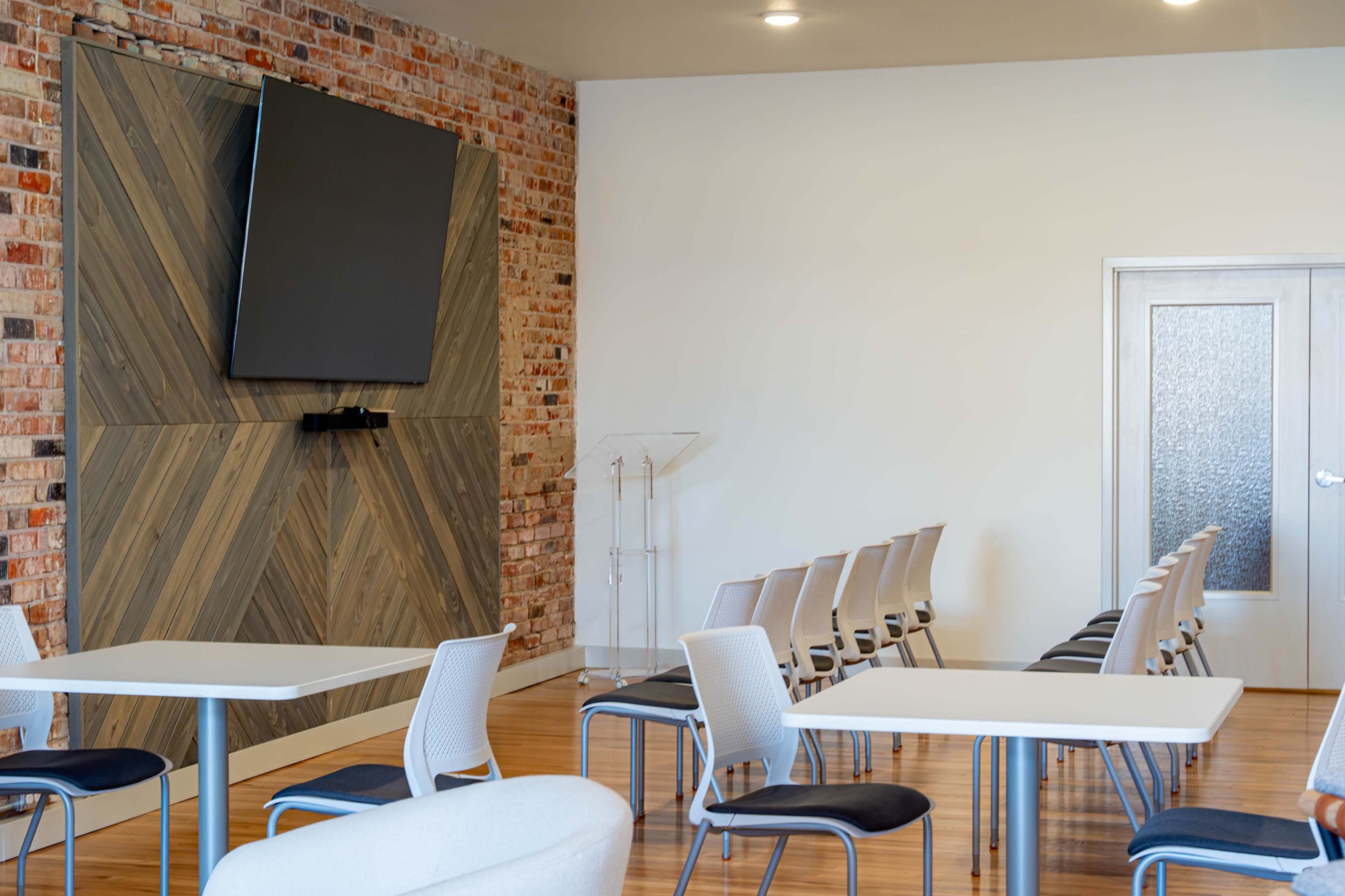 The image shows a modern meeting room featuring a brick accent wall, a large television mounted on a wooden panel, and rows of chairs arranged in front of a pair of tables.