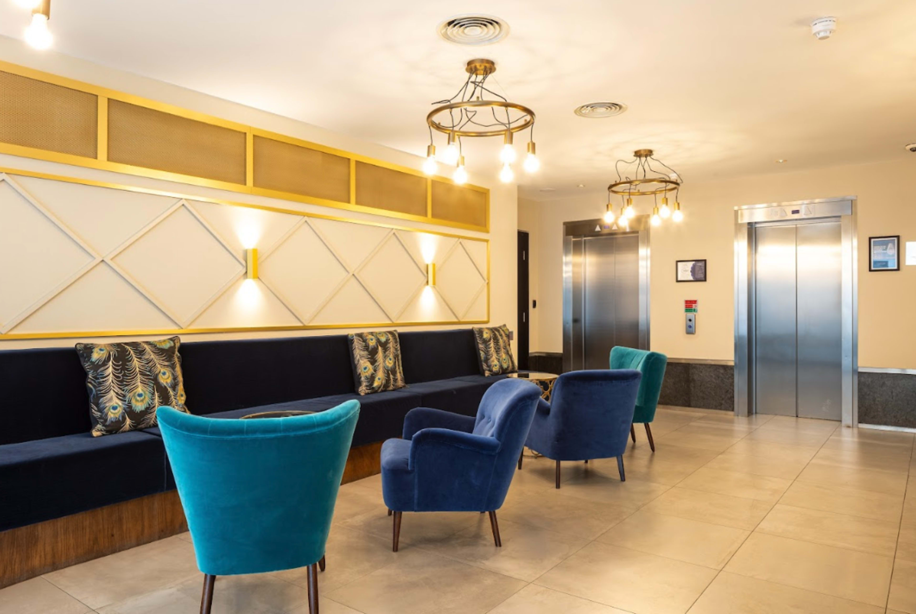 A modern lobby area with a row of blue chairs and a long dark bench against a light wall, featuring two elevators in the background.