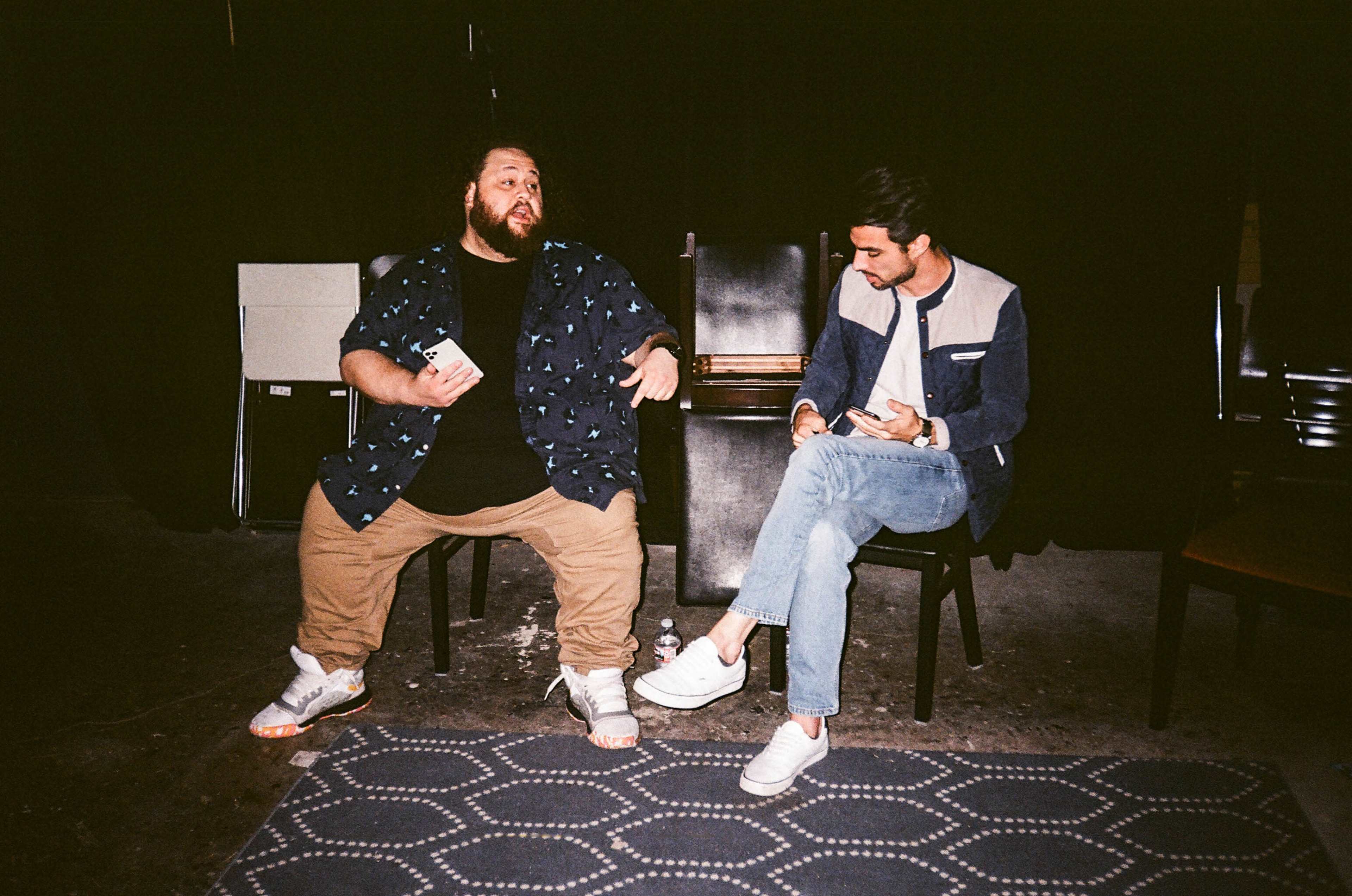 Two men sit on chairs in a dimly lit space, engaged in conversation, with one holding a small item and the other looking at his phone.