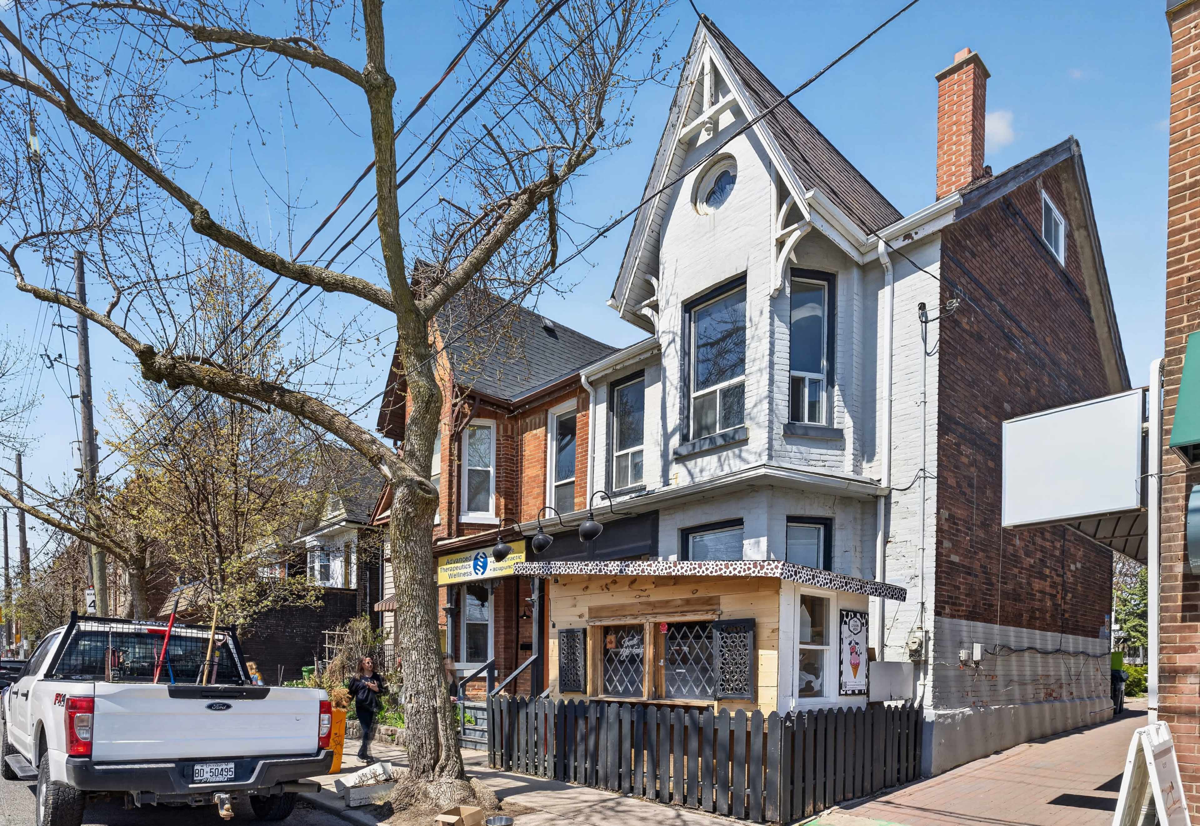 A two-story brick house with a gabled roof features a porch with a decorative railing and is situated on a tree-lined street.