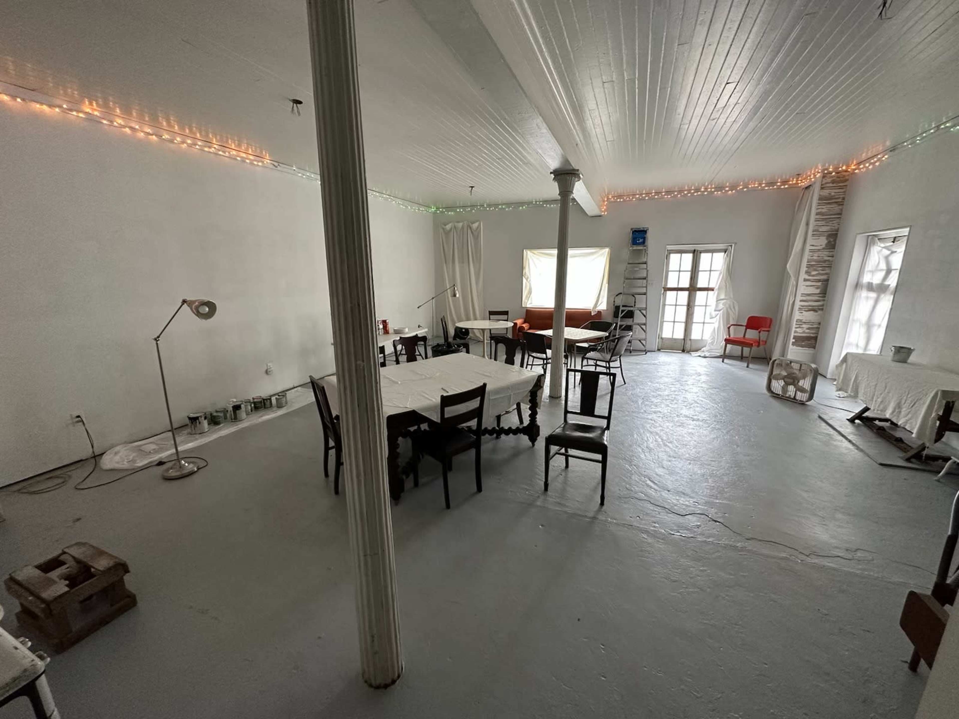 The image shows a spacious, uncluttered white room with several tables and chairs arranged, soft lighting along the walls, and large windows.