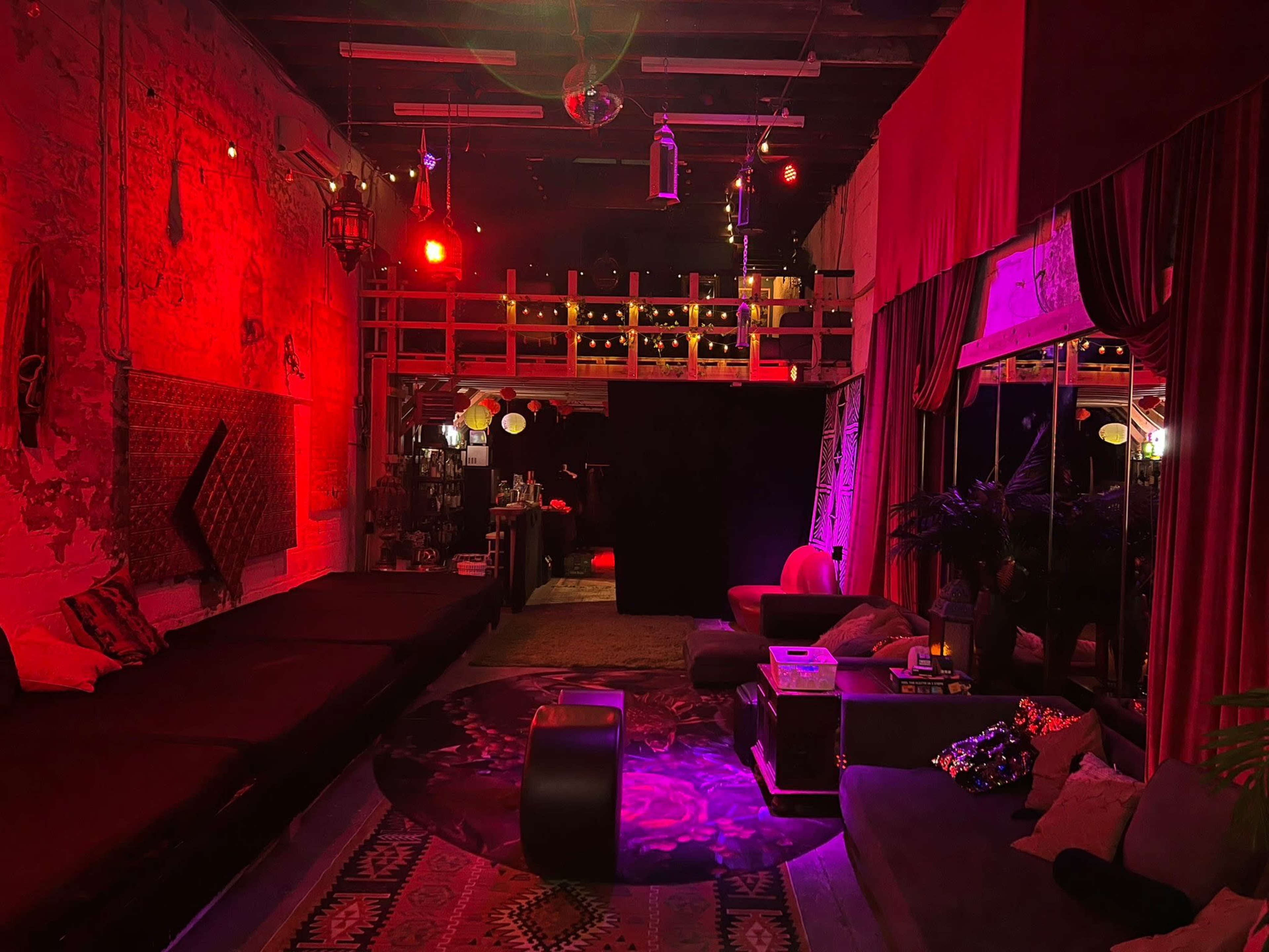 The image shows a dimly lit lounge space featuring red lighting, colorful decorations, and a mix of seating arrangements.