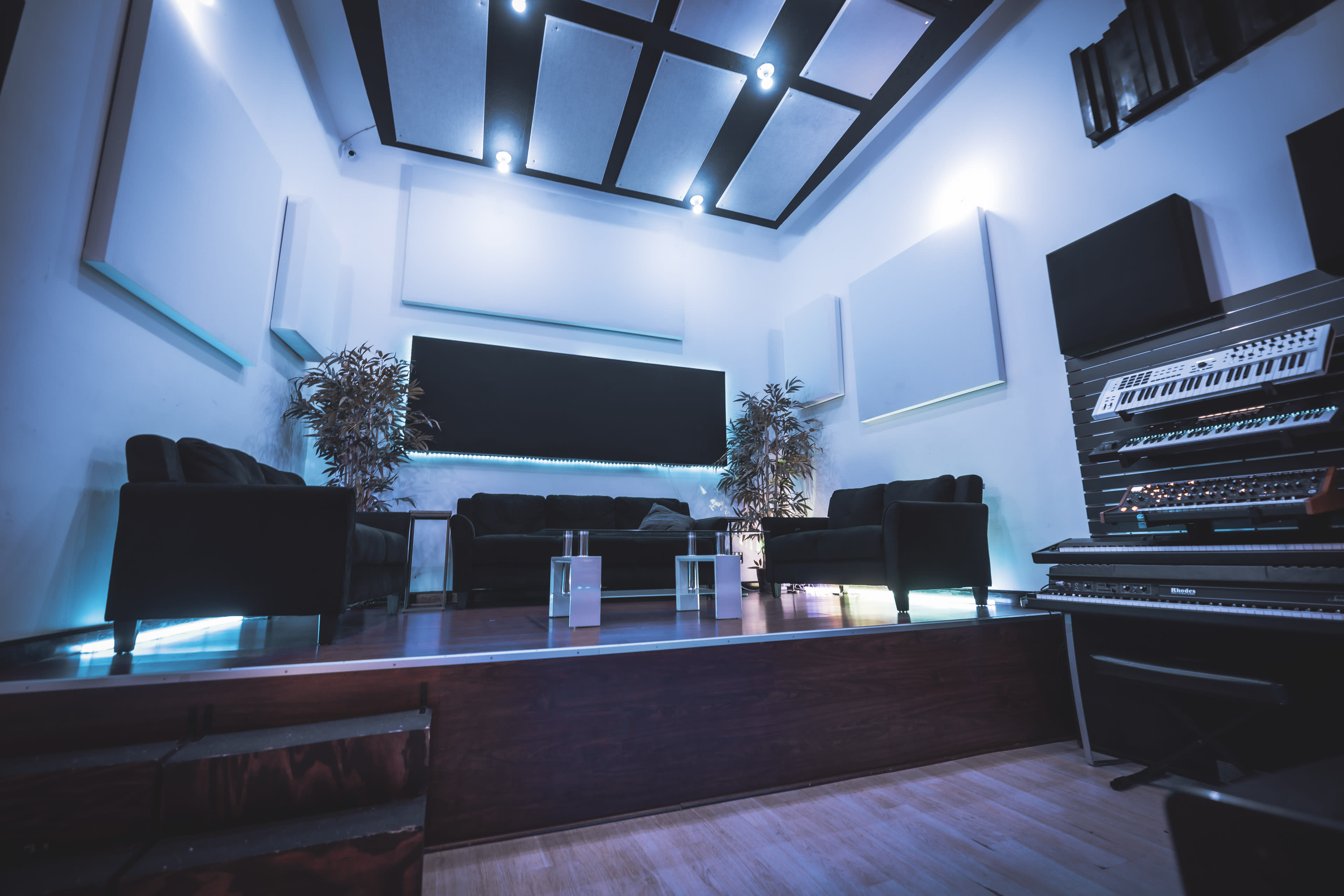 The image shows a modern studio lounge area with two black sofas, ambient lighting, plants, and electronic music equipment along one wall.