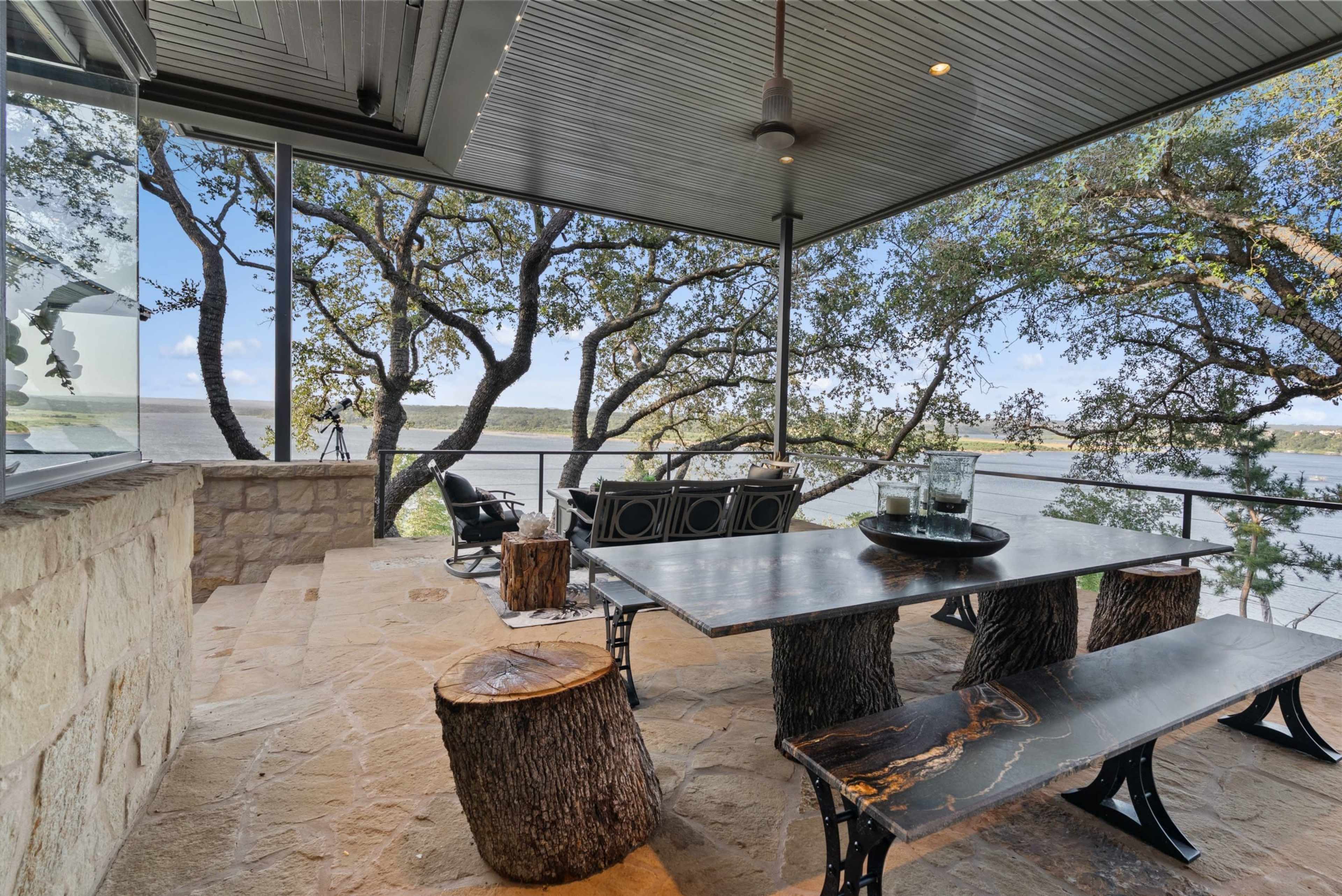 A covered outdoor dining area features a large wooden table surrounded by log-style stools, with a scenic view of a lake and trees in the background.