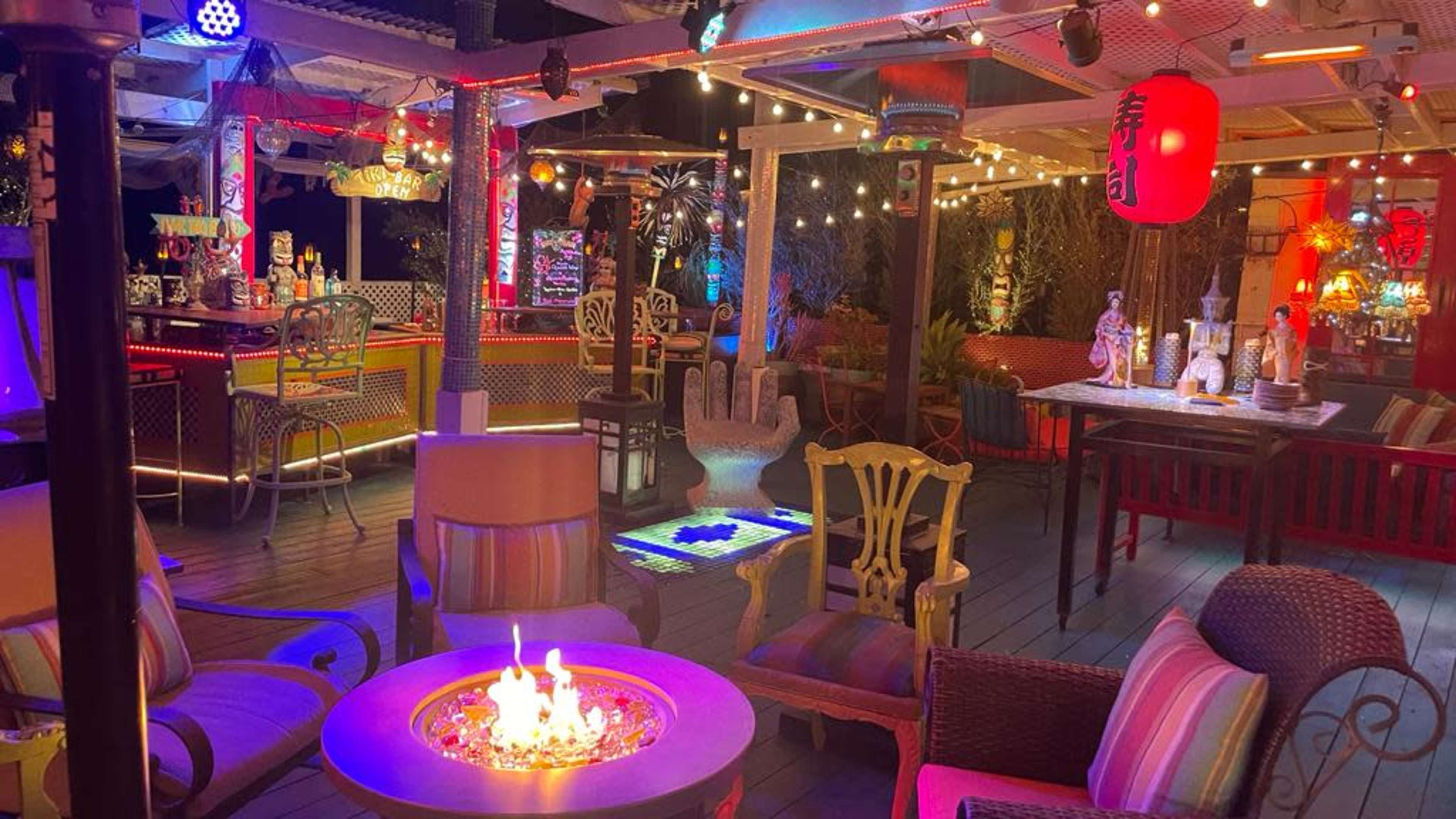 The image depicts a colorful outdoor lounge area with vibrant lighting, seating arrangements, and a central fire pit surrounded by decor typical of a tropical or festive setting.