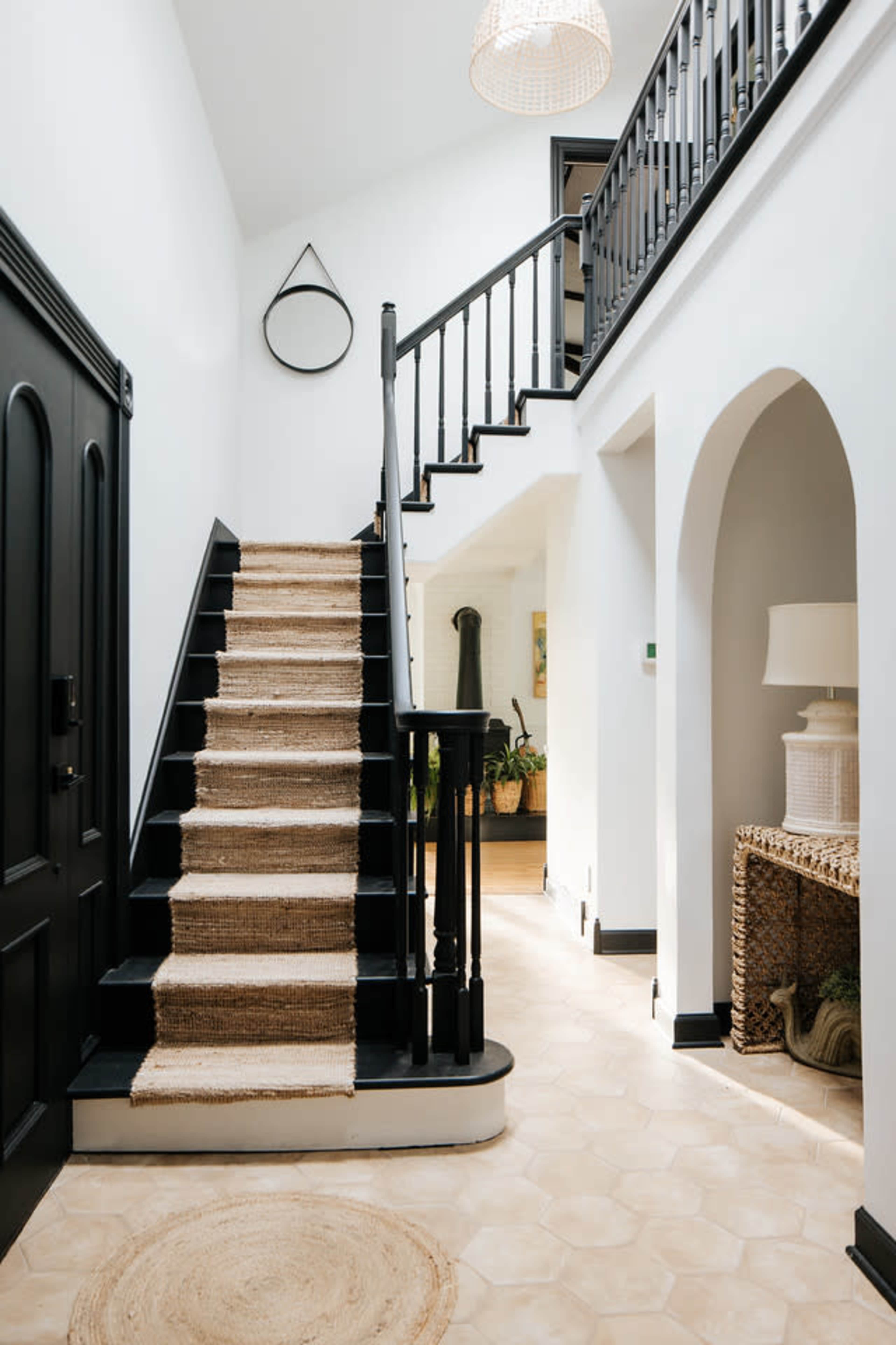 A well-lit hallway featuring a staircase with black railings and a natural fiber runner, leading to an upper level with light-colored walls and plants visible in the background.