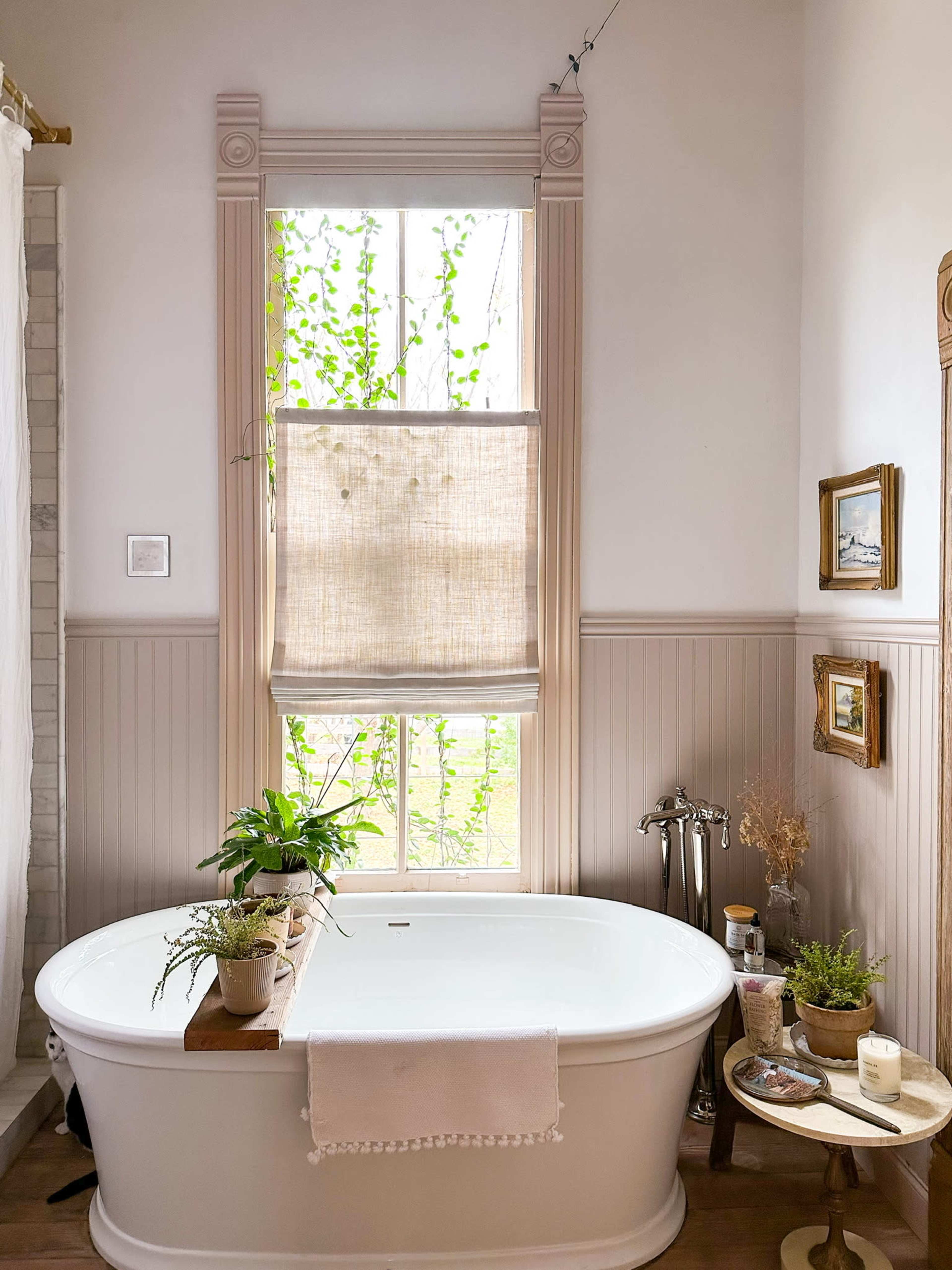 A spacious bathroom featuring a white freestanding bathtub, a window with greenery outside, and various plants placed around the room.