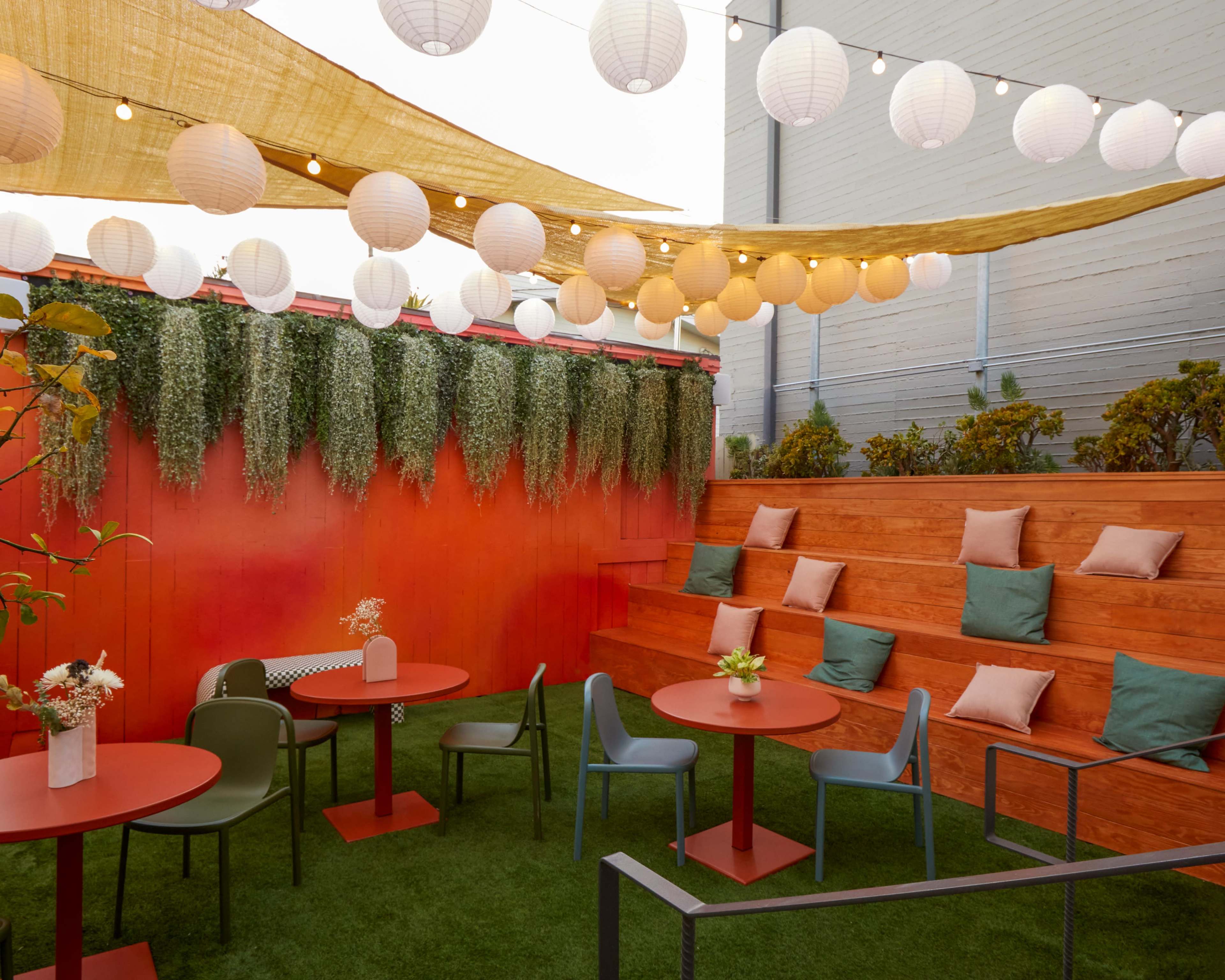 A vibrant outdoor seating area features wooden steps, round tables, and lanterns overhead, surrounded by greenery and decorative pillows.