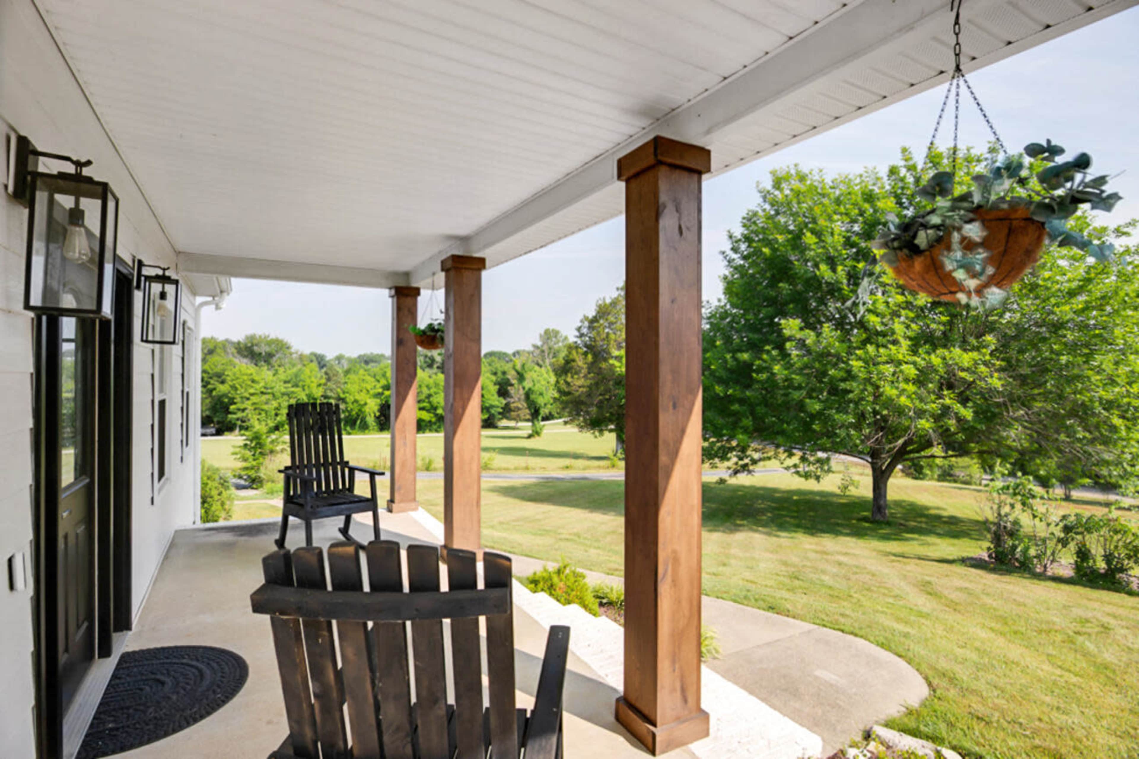 A front porch with two rocking chairs, wooden columns, and hanging planters, overlooking a green lawn and trees.