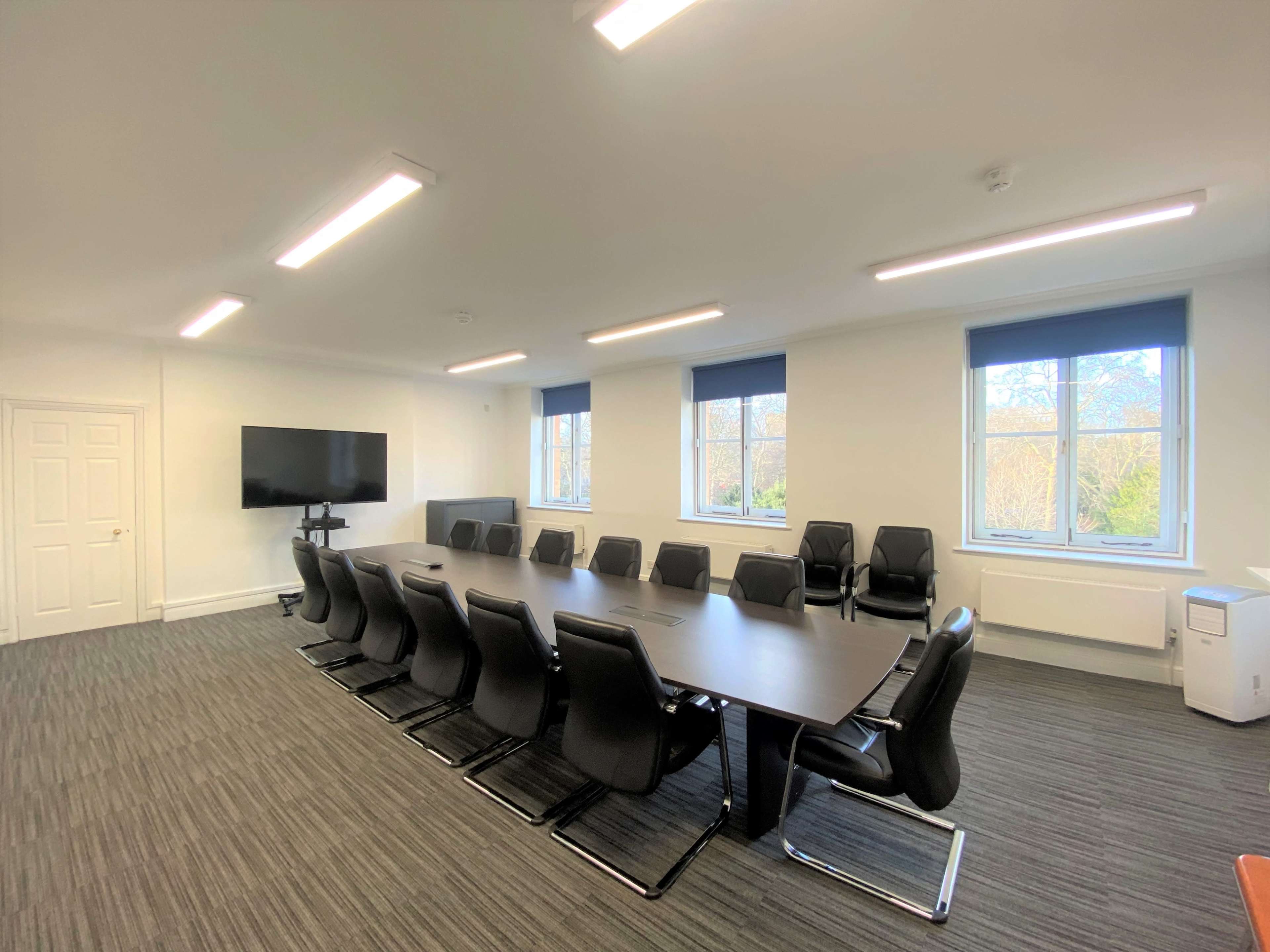 The image shows a modern conference room with a long table, black chairs, a large screen on the wall, and windows providing natural light.