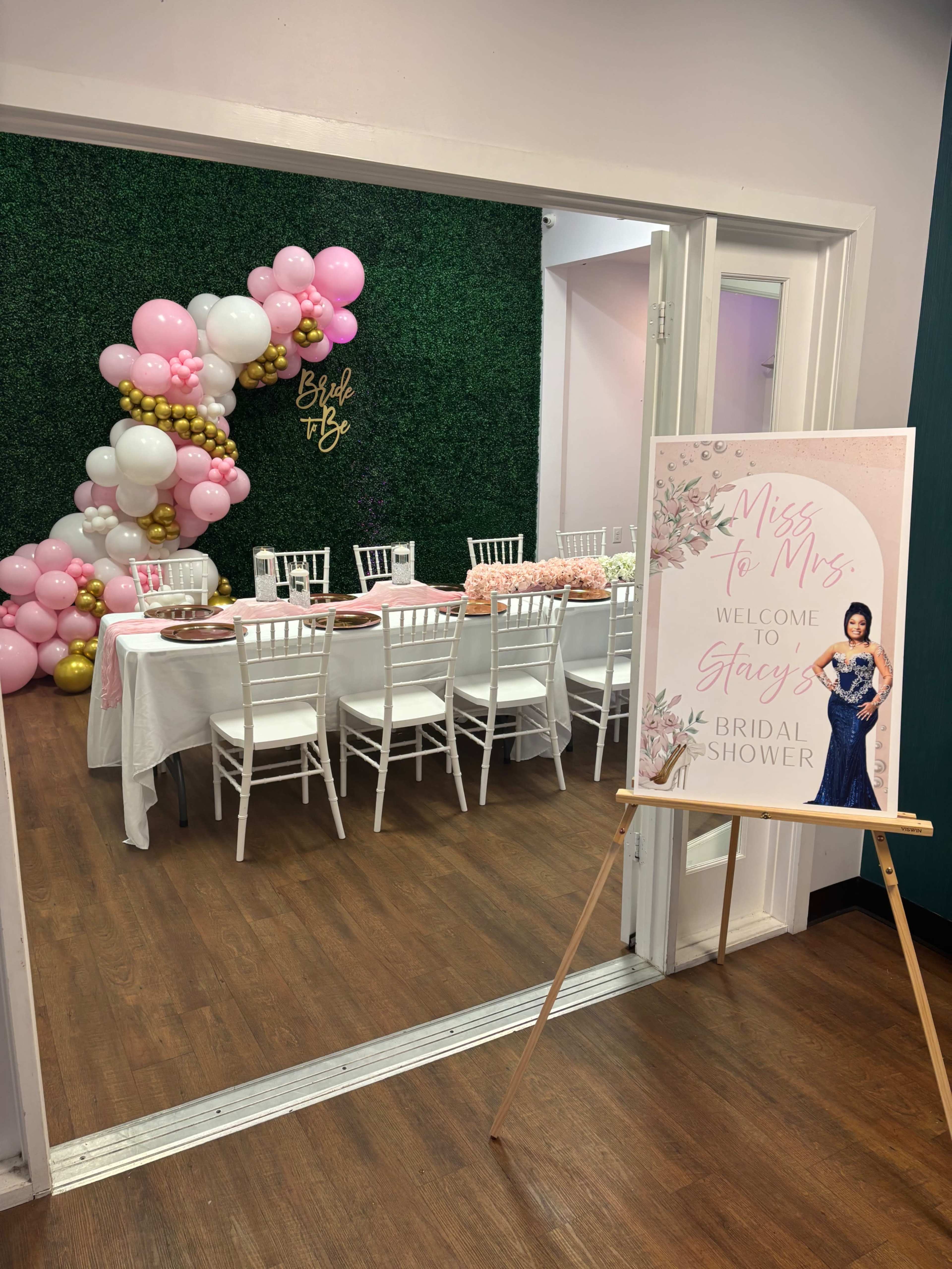 The image shows a decorated room set up for a bridal shower, featuring a table with food and drinks, white chairs, and a balloon arch in pink and gold hues.