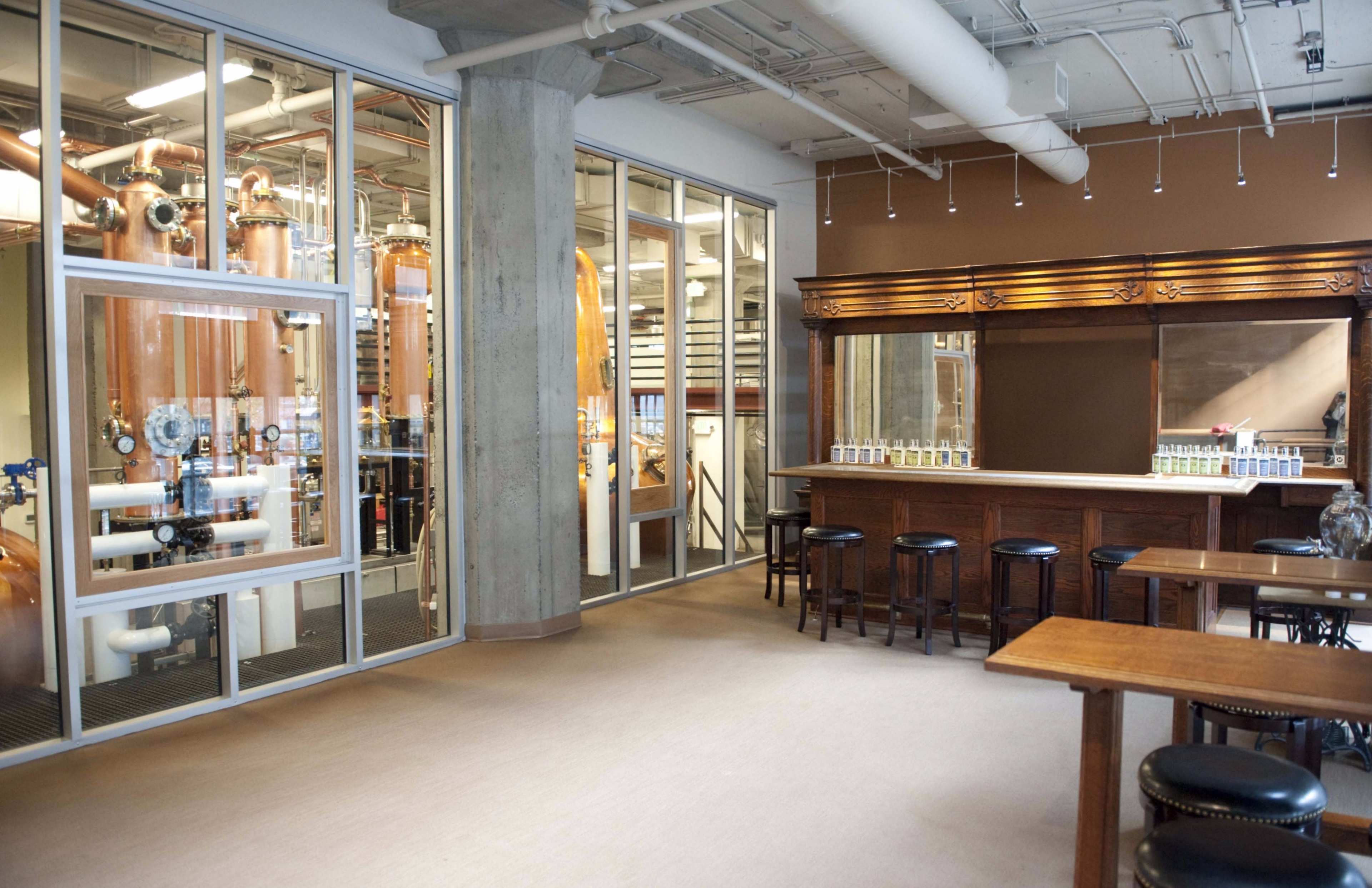 A spacious industrial-style room with large glass windows revealing copper distillation equipment, paired with a wooden bar and several stools in the foreground.