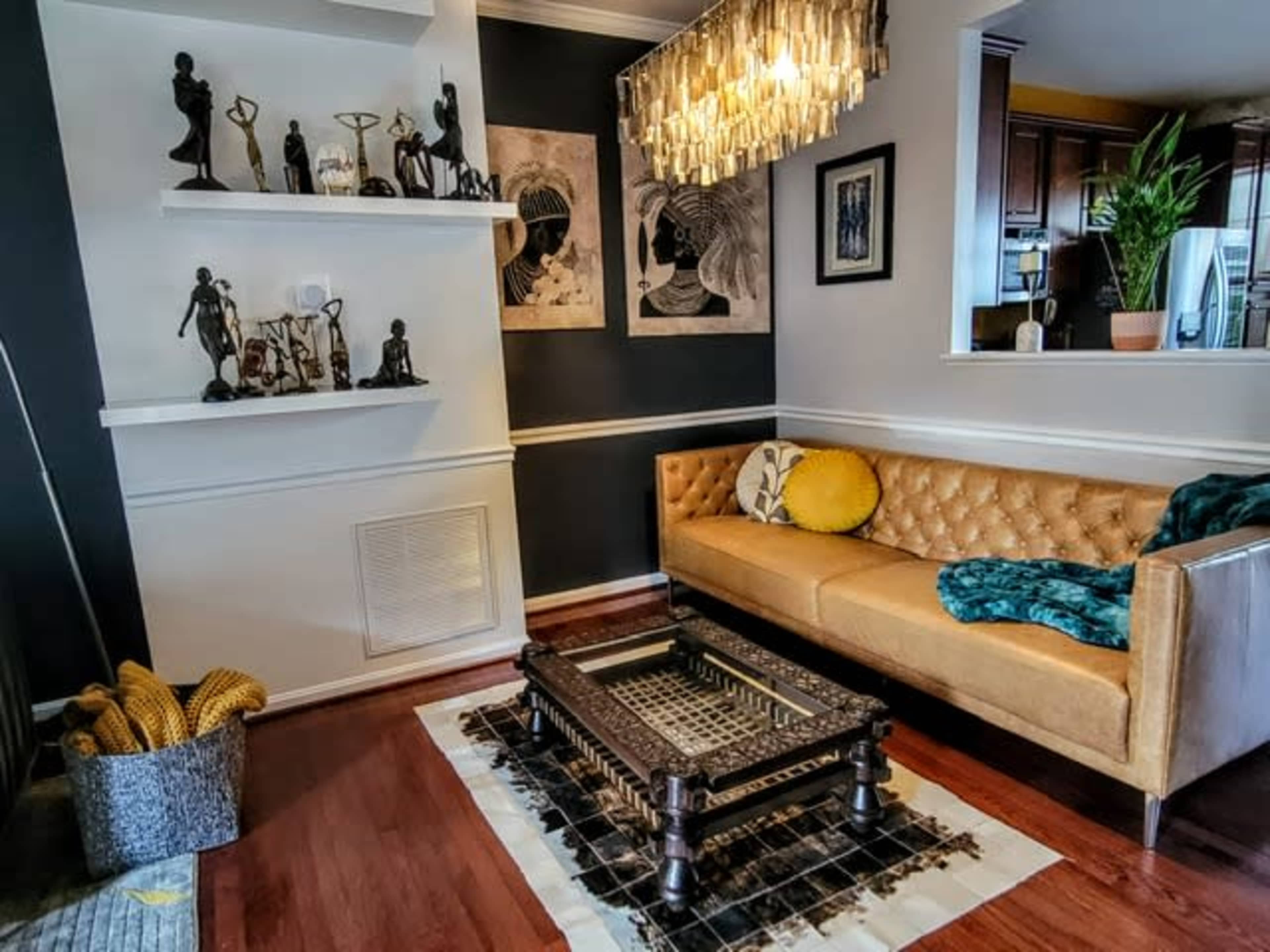 A living room with a tan tufted sofa, decorative sculptures on shelves, and a glass chandelier above a low, ornate coffee table.