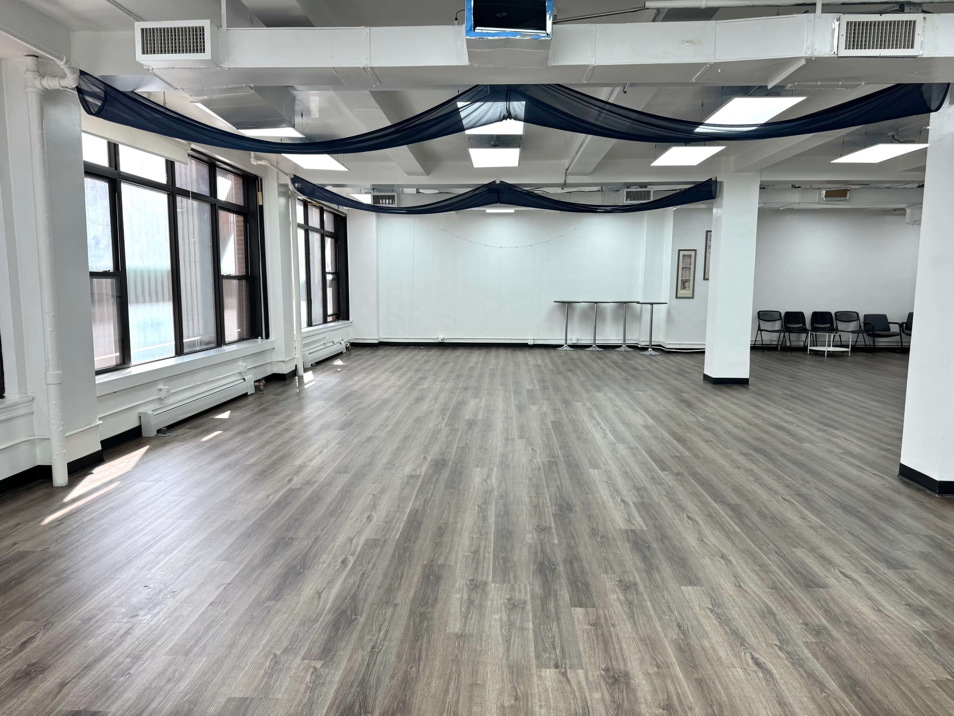 The image shows an empty, well-lit interior space with a wooden floor, large windows, and simple furniture in the background.