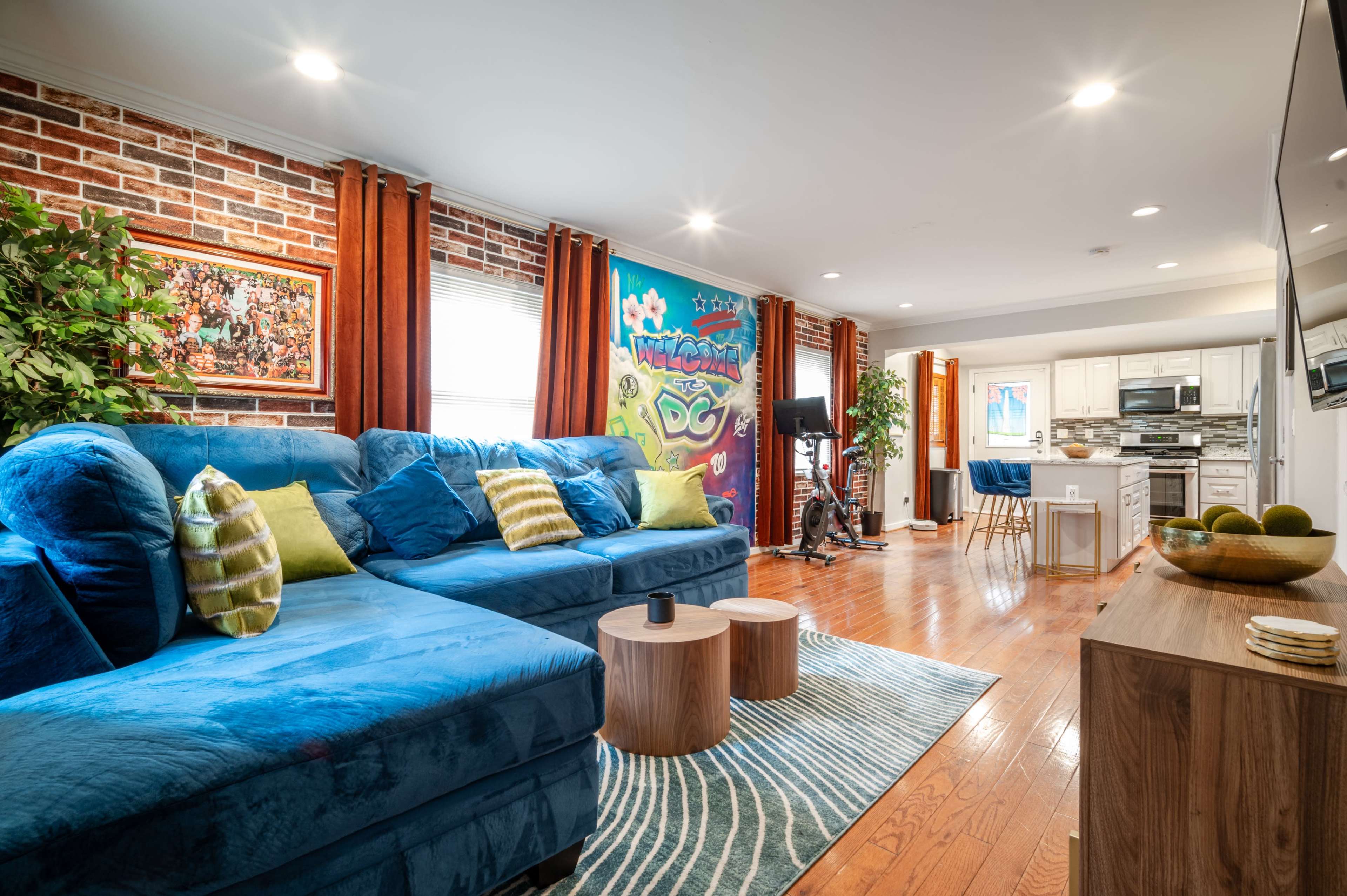 A cozy living room features a blue sectional sofa, wooden coffee tables, and a vibrant wall tapestry, leading into a well-lit kitchen area.