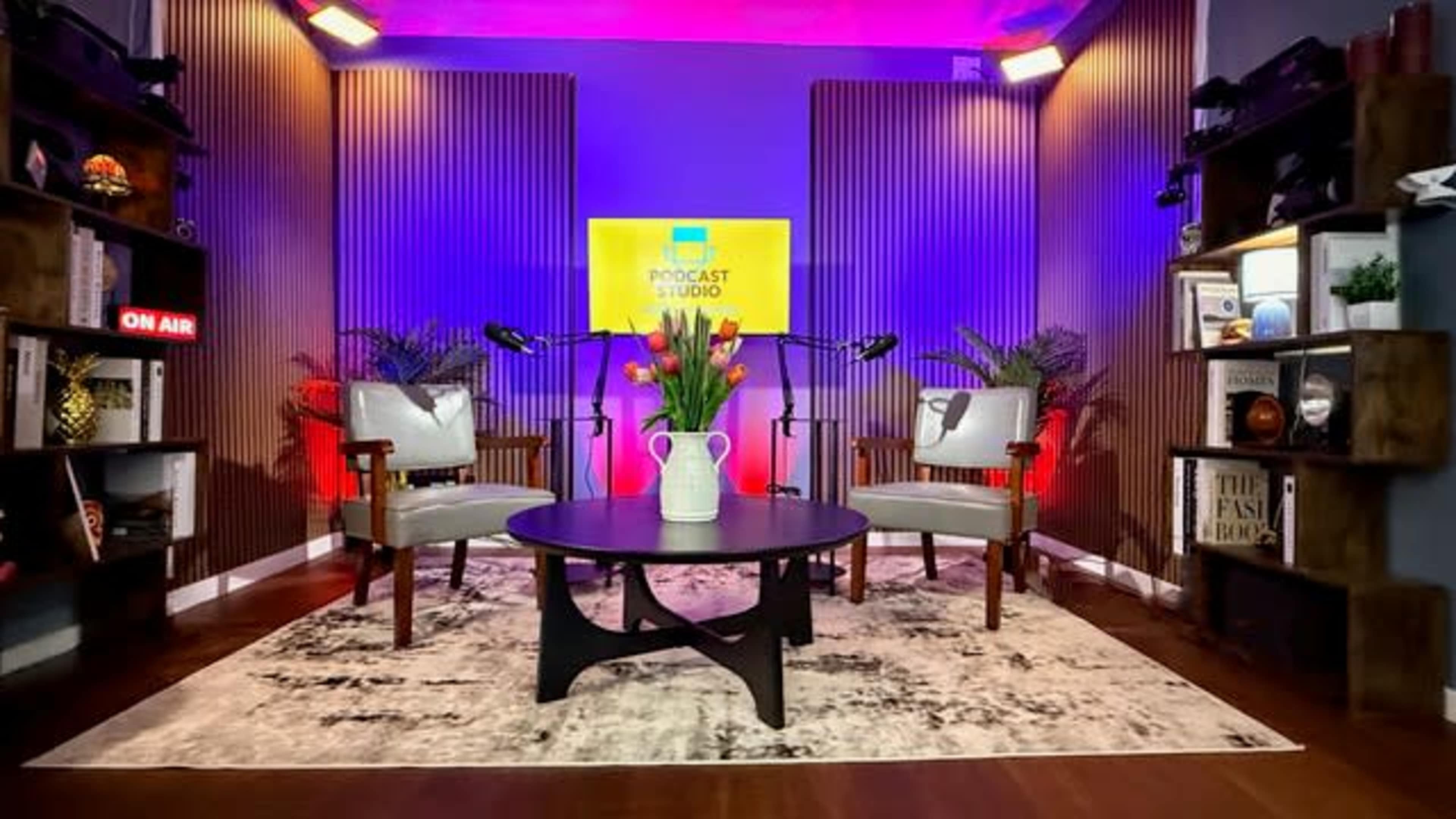 The image shows a modern podcast studio featuring two chairs, a round table, and shelves with decorations, all illuminated with colorful lighting.