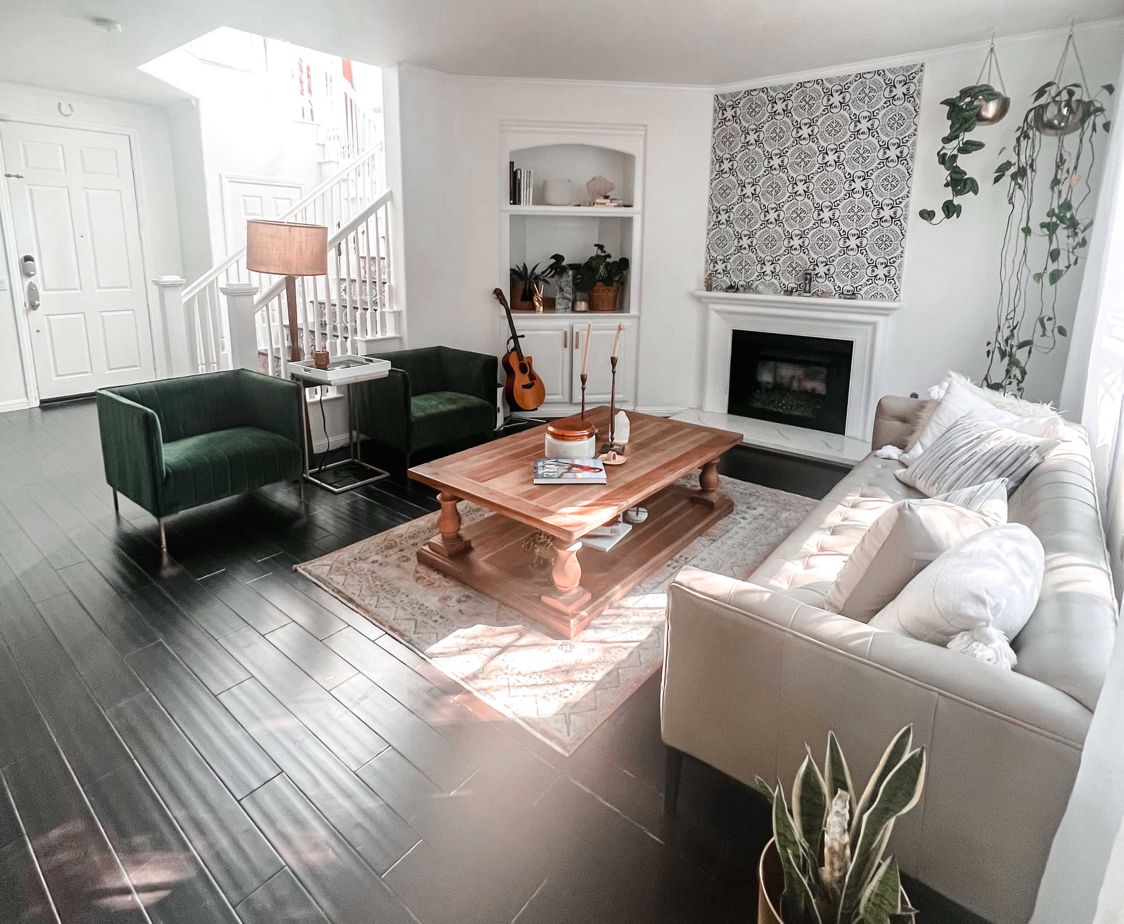 The image shows a bright living room with a cream sofa, green armchairs, a wooden coffee table, and a guitar placed near a white fireplace.