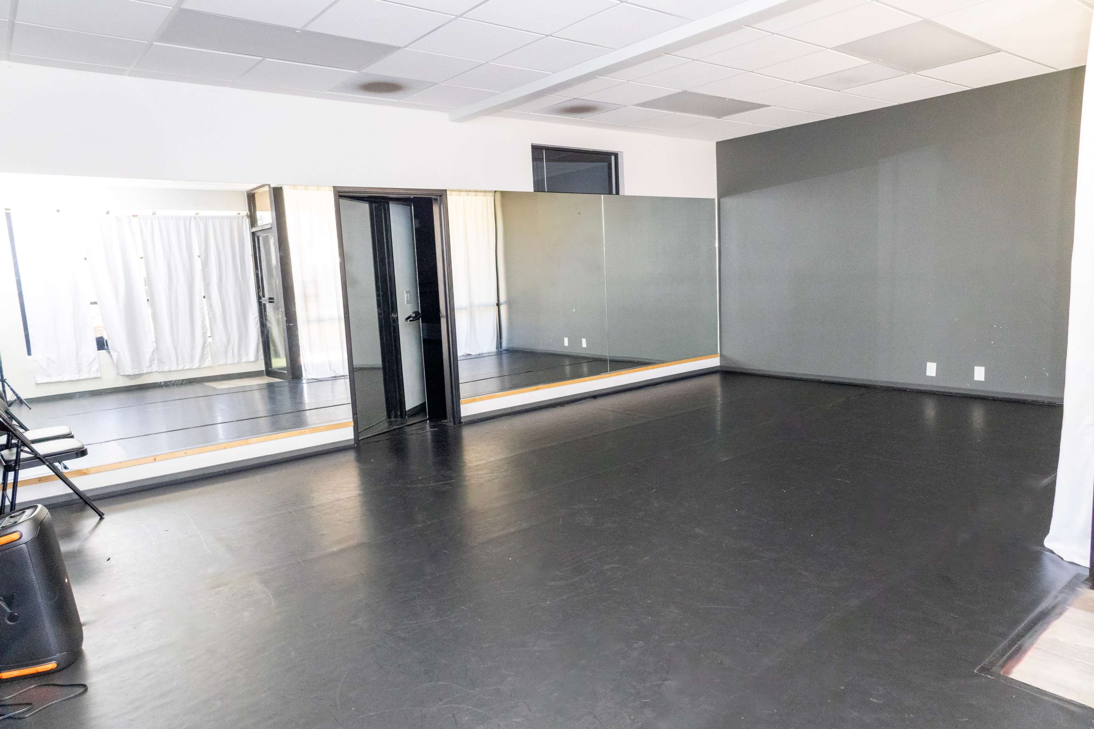 The image shows an empty dance studio featuring a mirrored wall, black flooring, and several chairs lined up against the side.
