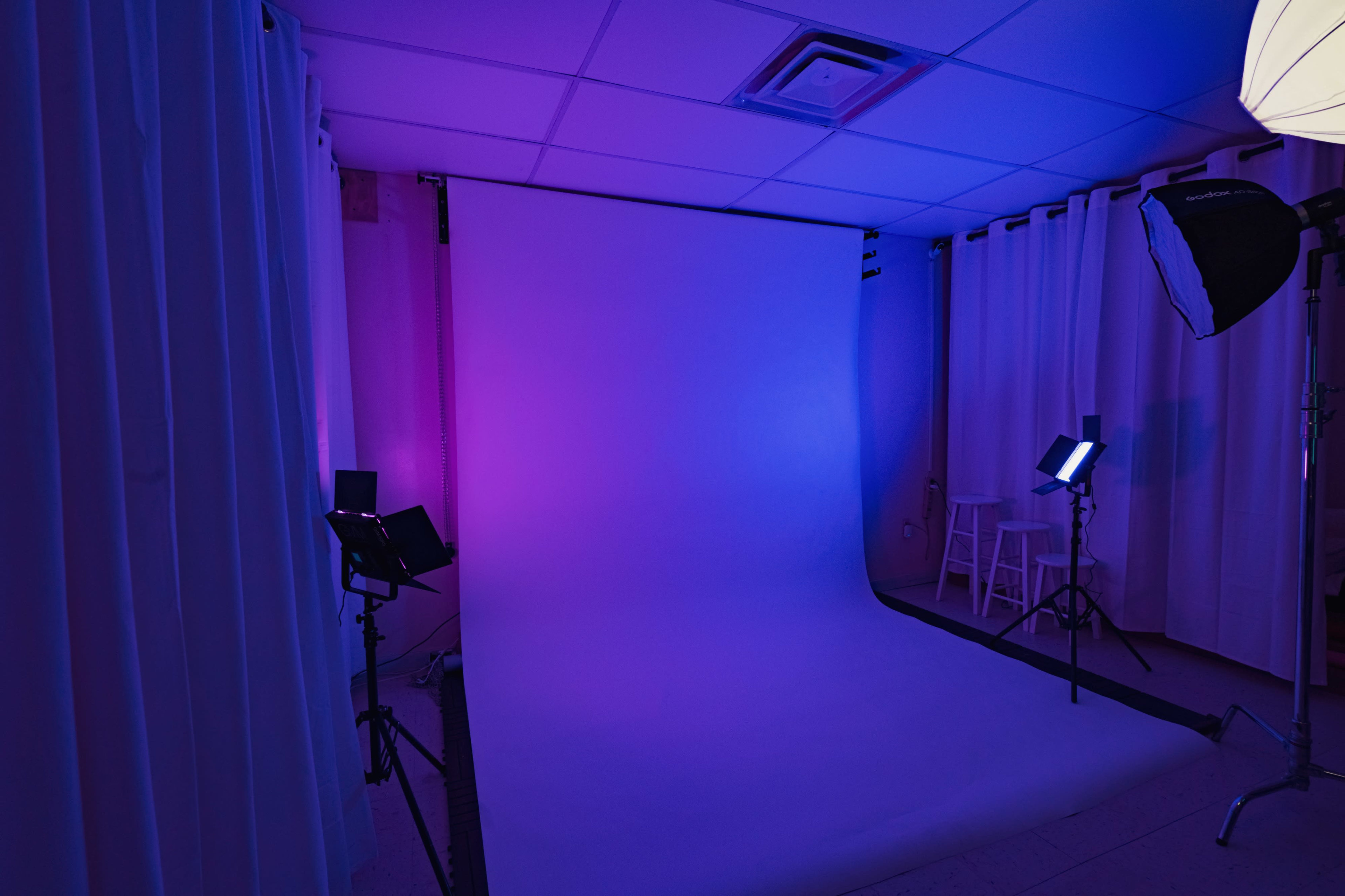 A photography studio features a backdrop with varying shades of purple and blue lighting, flanked by softboxes and stands for lighting equipment.