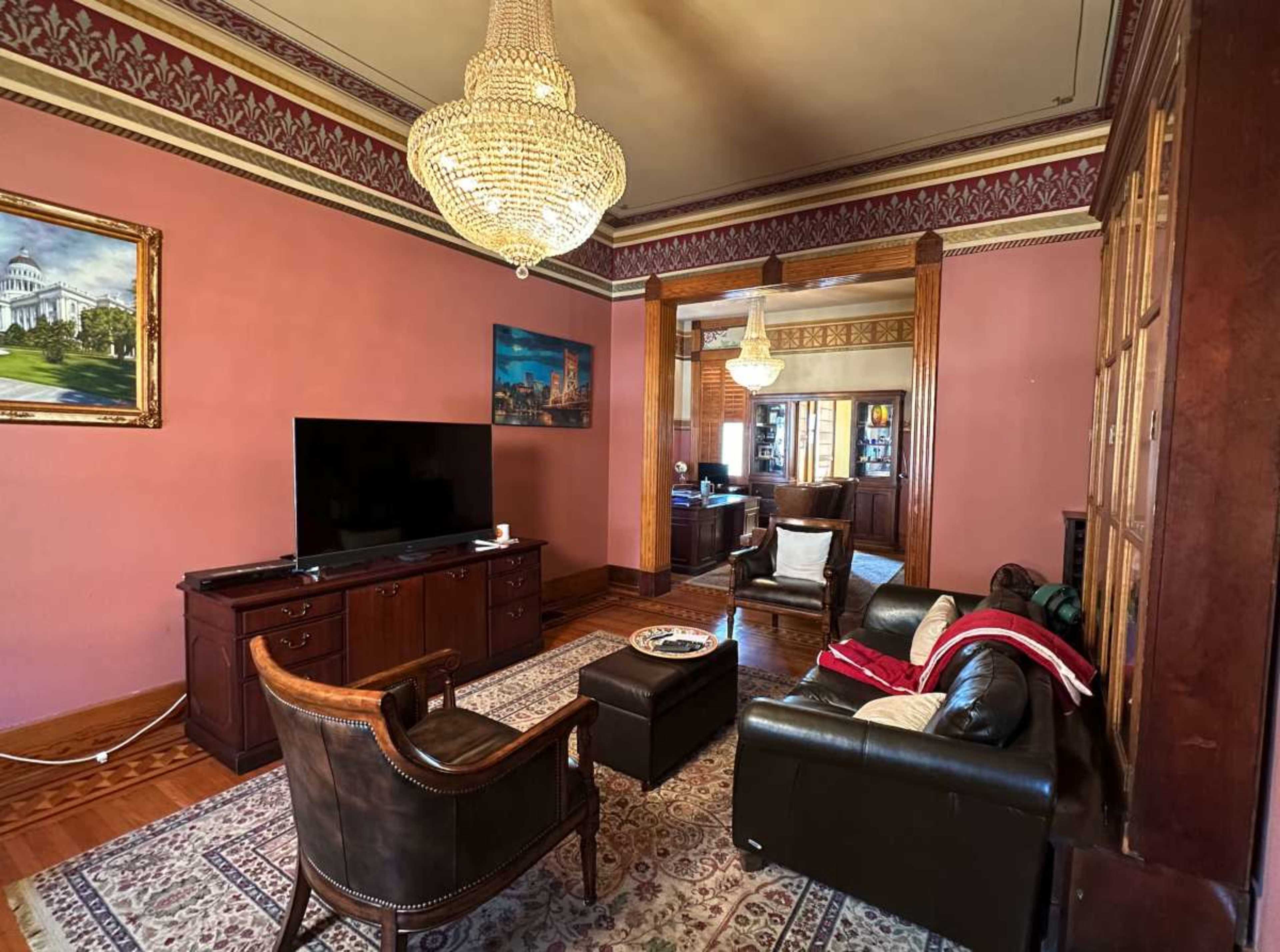 The image shows a vintage living room with a chandelier, two leather sofas, a television, and decorative wall details.