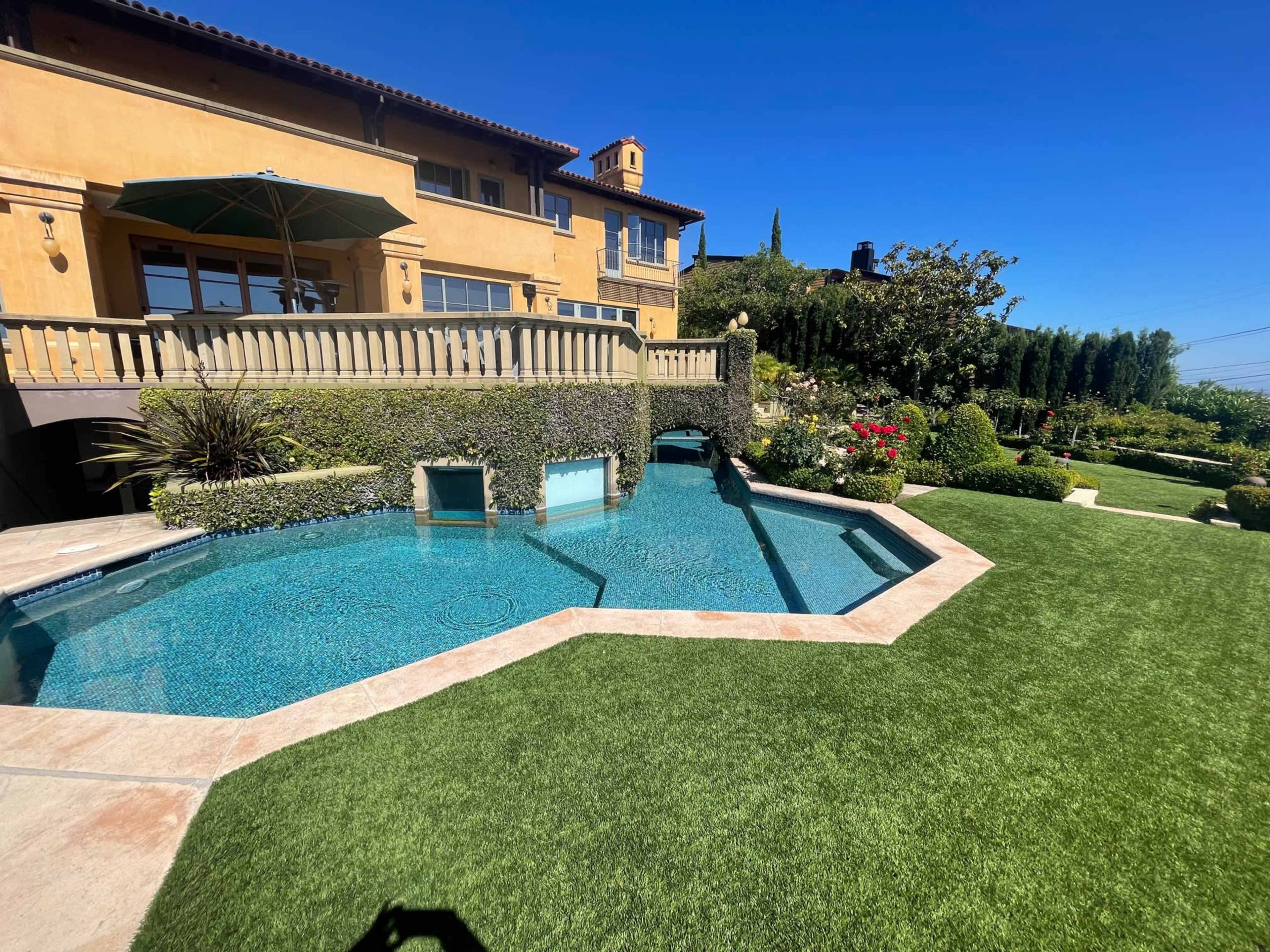 The image shows a landscaped backyard with a modern swimming pool, trimmed hedges, and a yellow stucco house under a clear blue sky.