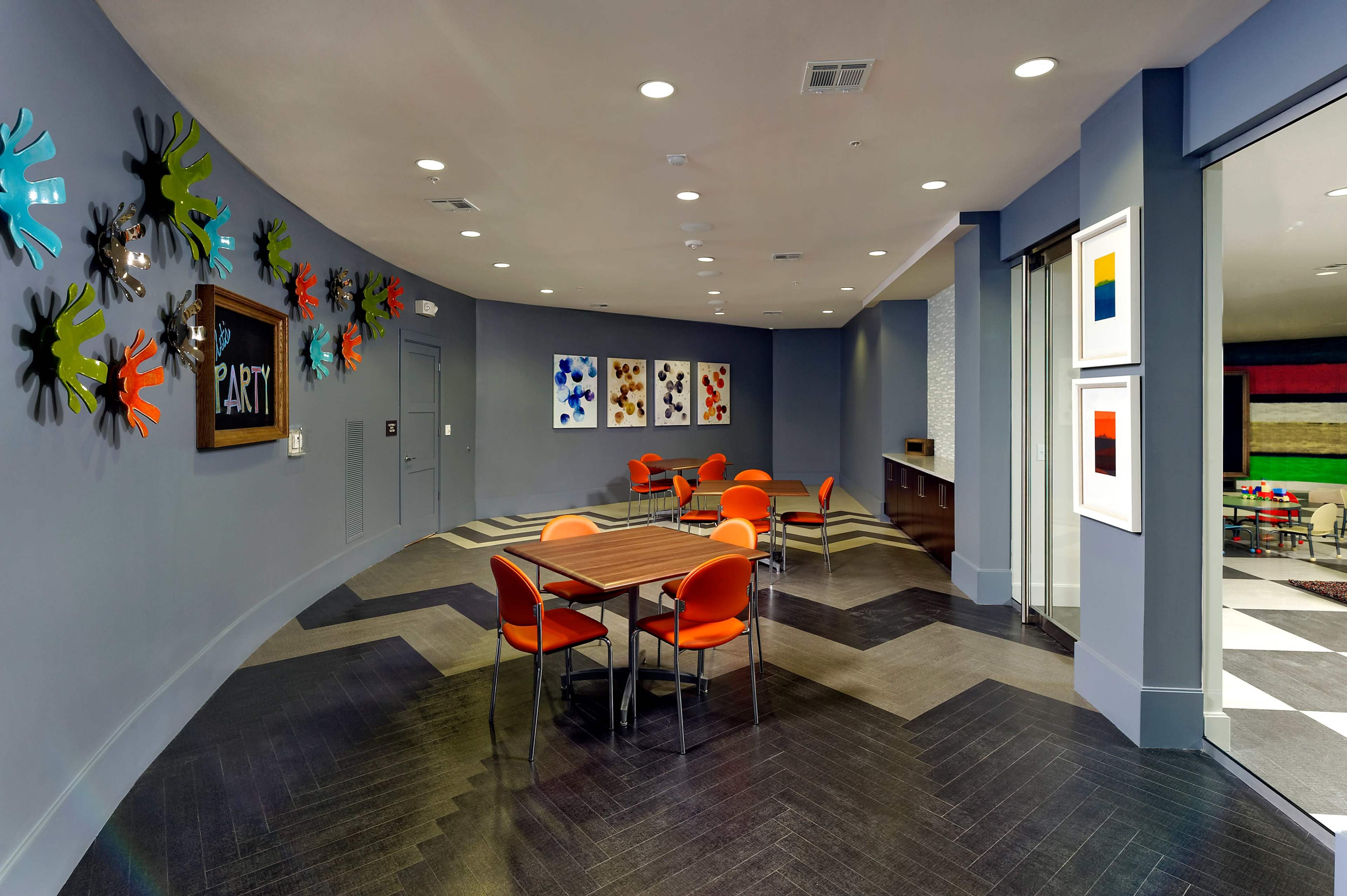 A circular lounge area features round tables with orange chairs, walls adorned with colorful artwork, and handprint decorations.