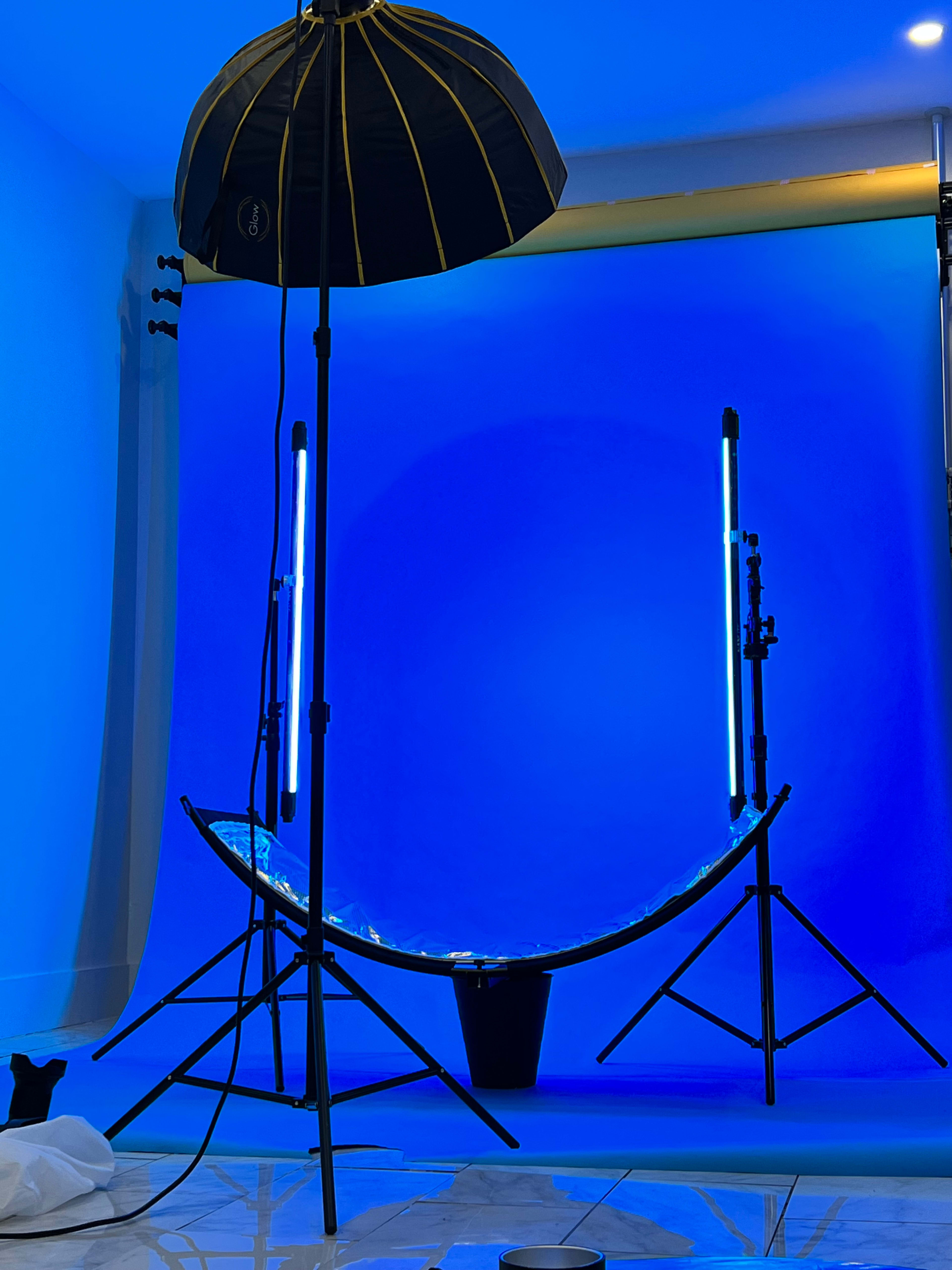 A photography setup featuring a blue backdrop, a large umbrella light, and two vertical light stands arranged on a tiled floor.