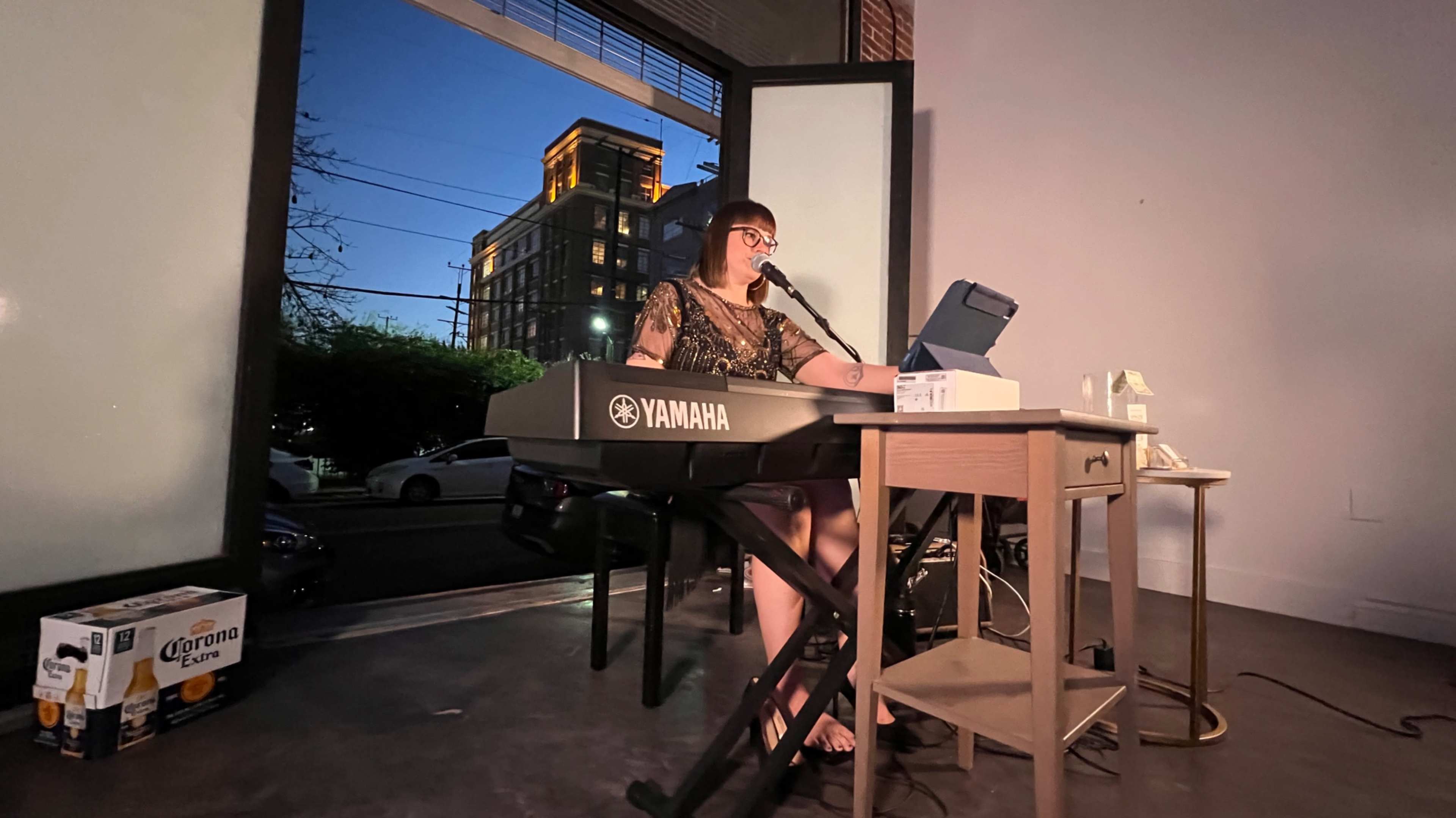 A woman plays a keyboard and sings at a small table inside a venue, with city buildings visible through large windows behind her.