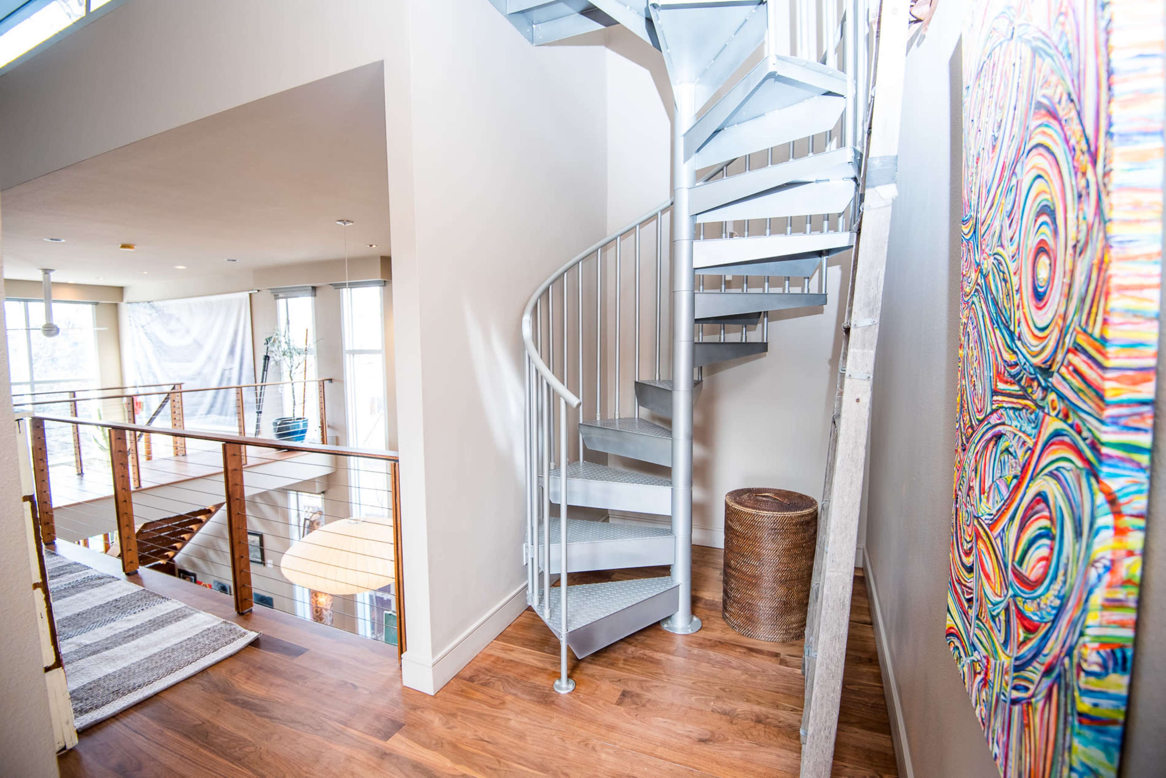 A spiral staircase made of metal leads to an upper level in a modern interior with wooden floors and colorful artwork on the wall.