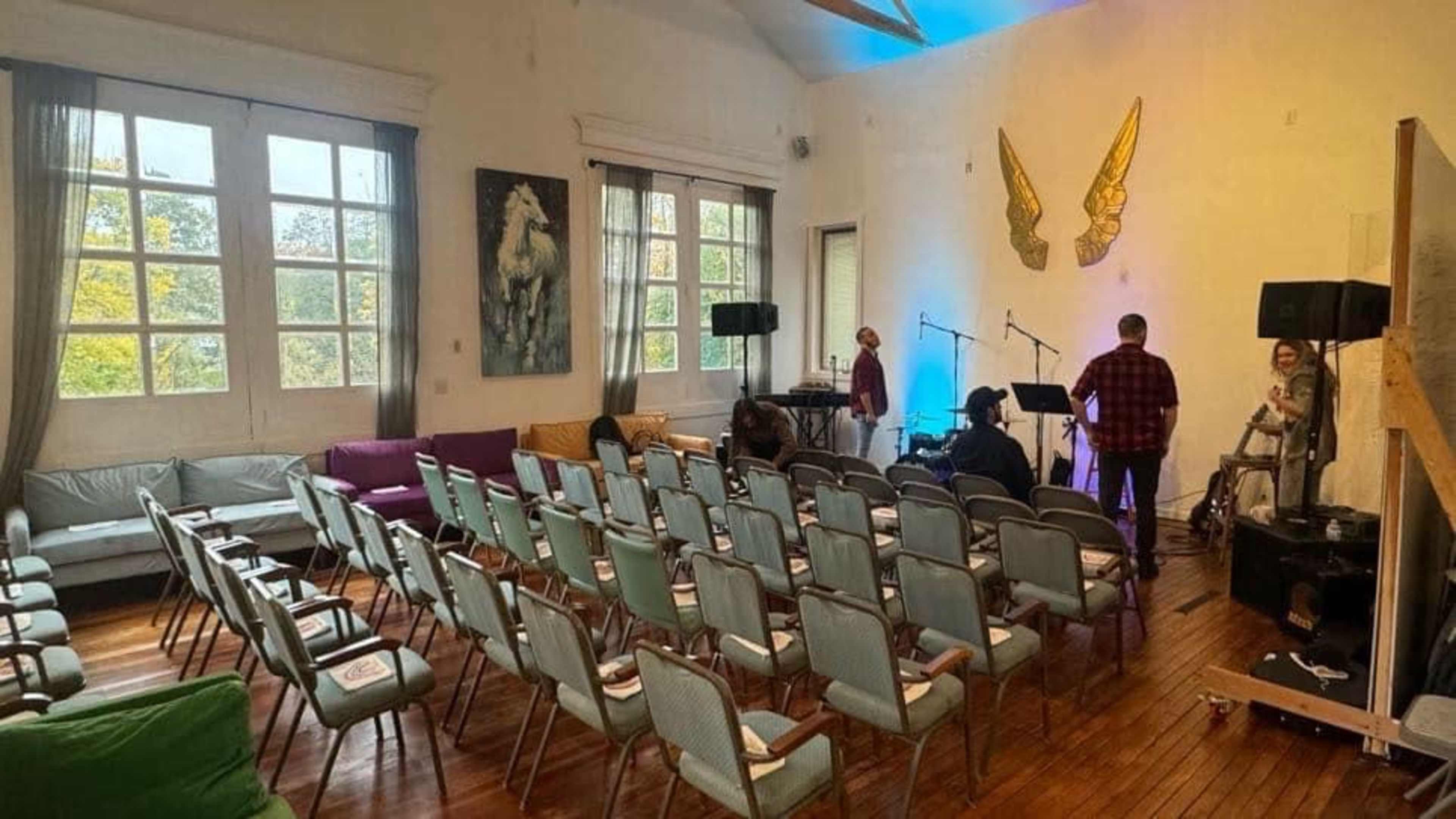 The image shows a spacious room with rows of empty chairs, a small stage set up for a performance, and large windows allowing natural light to fill the space.