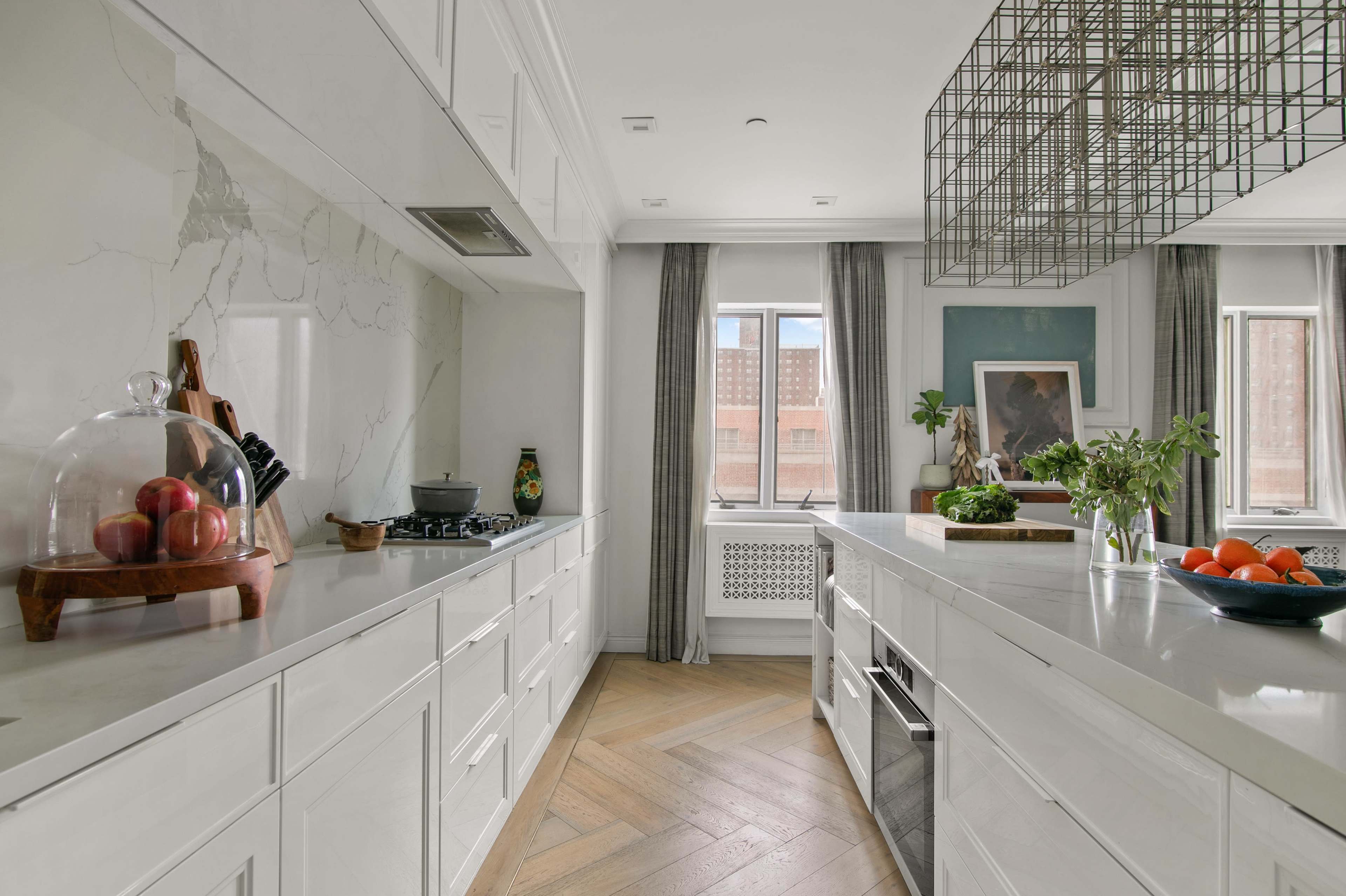 The image depicts a modern kitchen with white cabinetry, a central island, and a large window letting in natural light, featuring decorative items and fresh produce on the counters.
