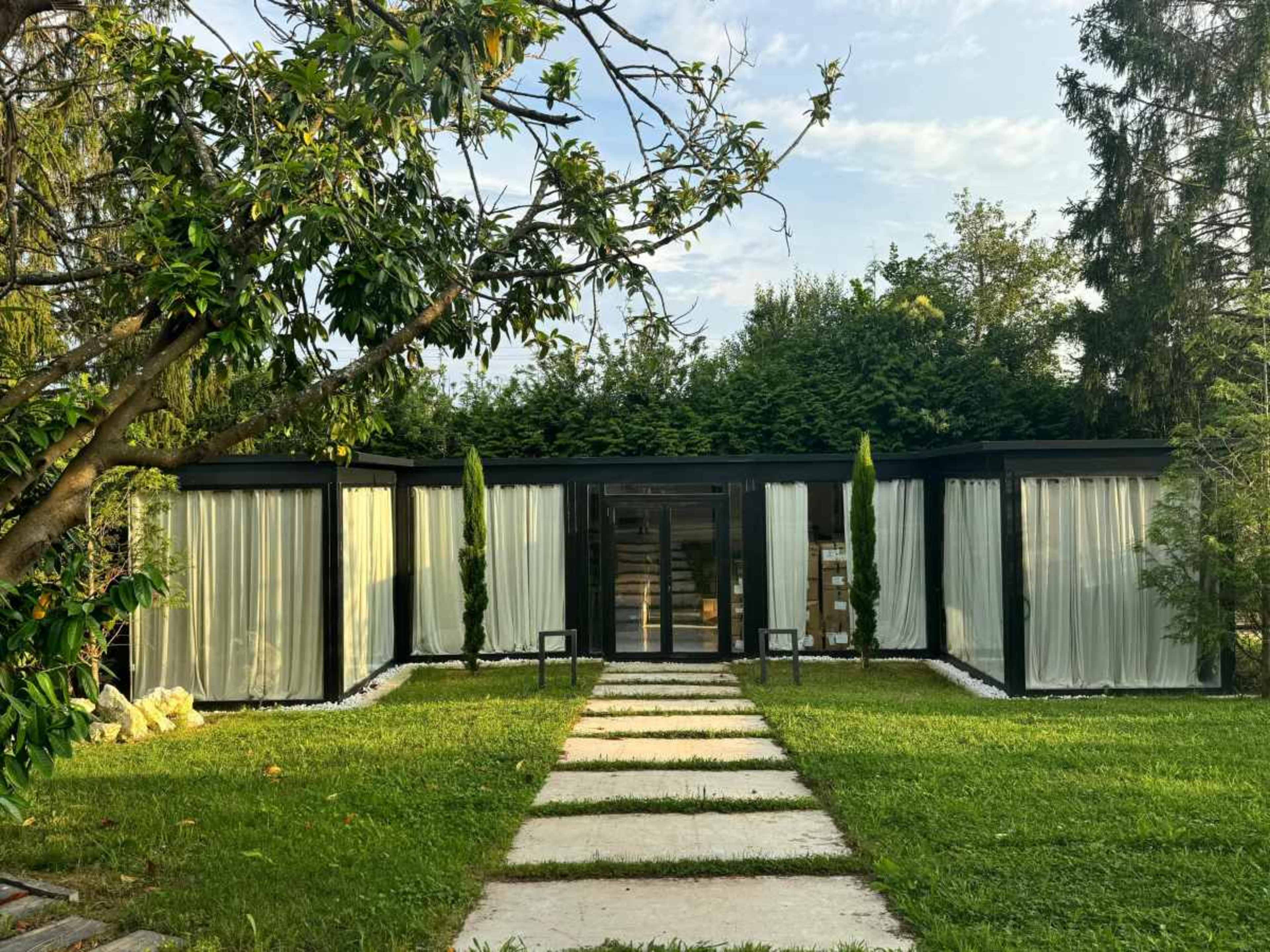 A modern, single-story building with large glass windows is surrounded by greenery and features a pathway leading to its entrance.