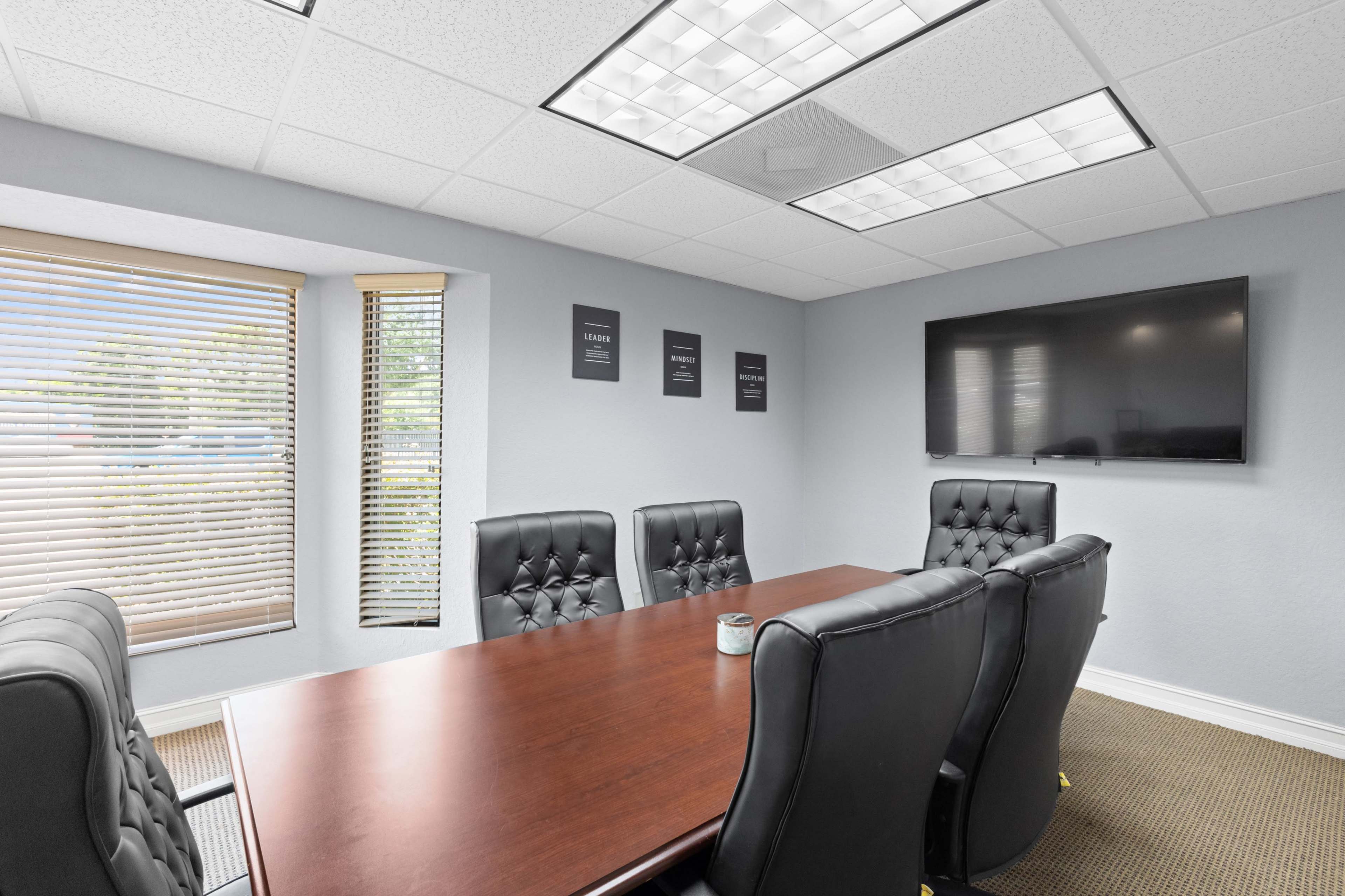 A conference room features a wooden table surrounded by black leather chairs, with blinds covering the windows and a wall-mounted television.