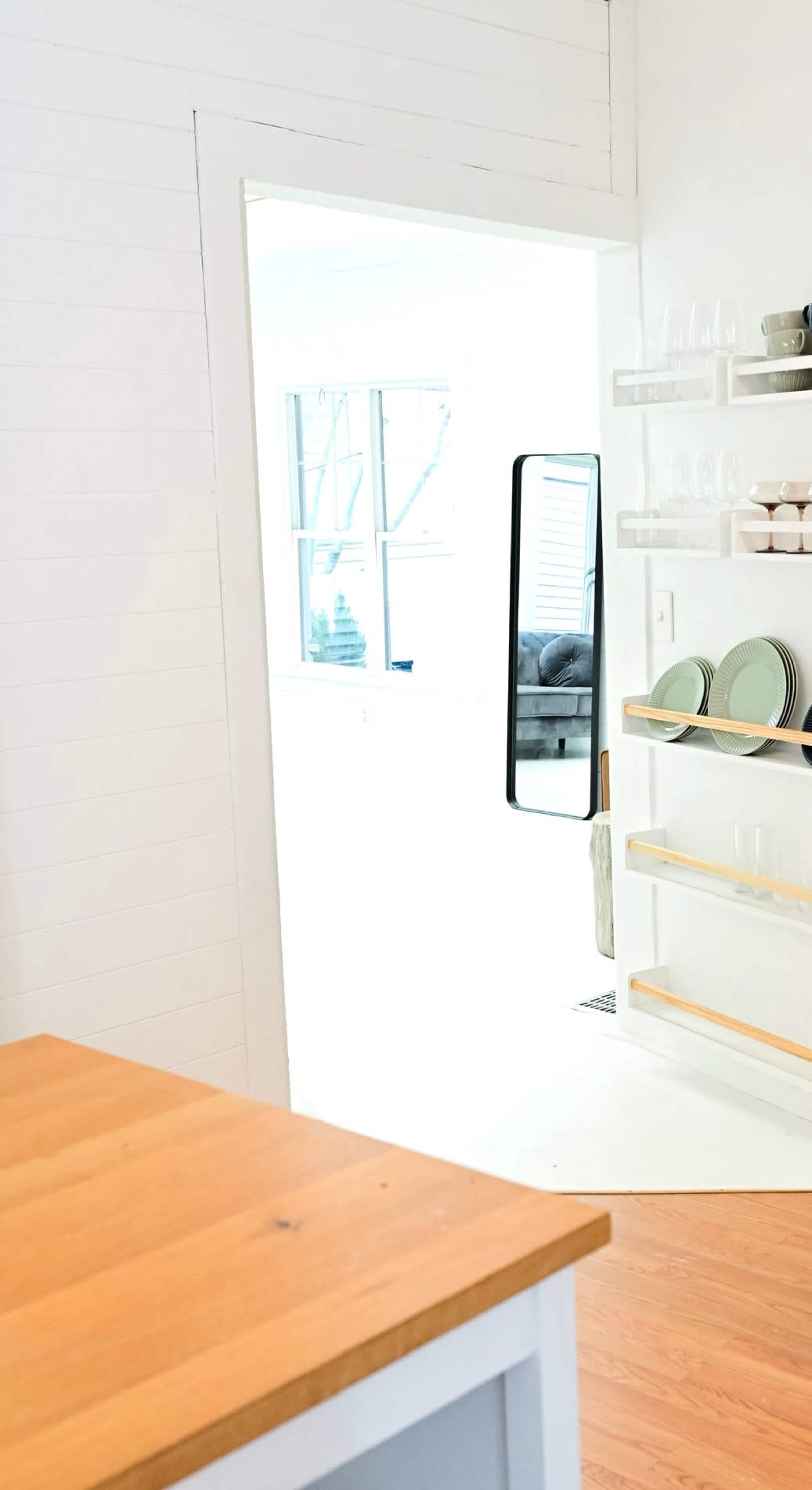 A bright, minimalistic kitchen features a wooden countertop, a mirror reflecting a neighboring room, and open shelving displaying dishware.