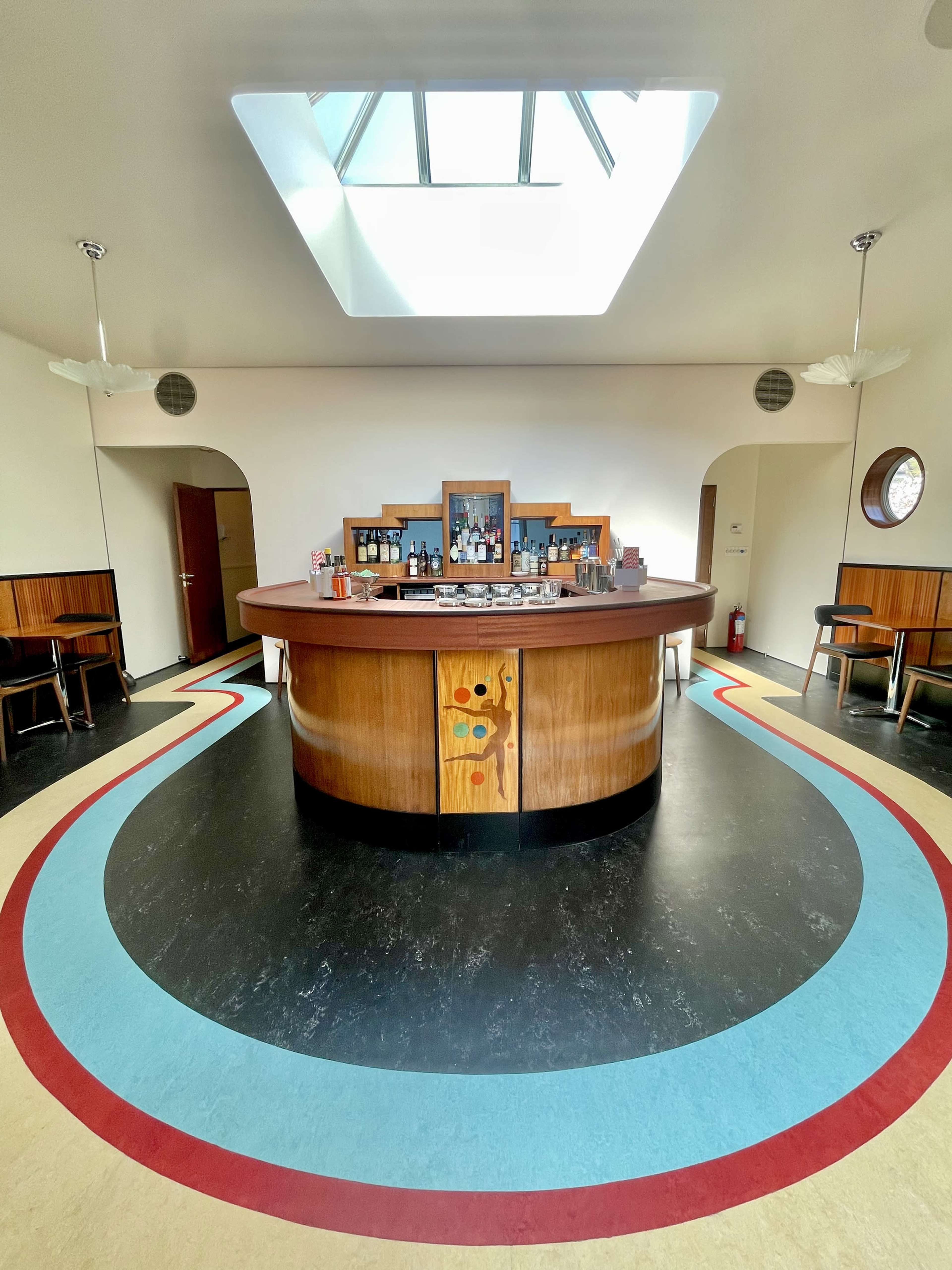 A circular wooden bar in a spacious interior with colorful curved flooring and a skylight above.
