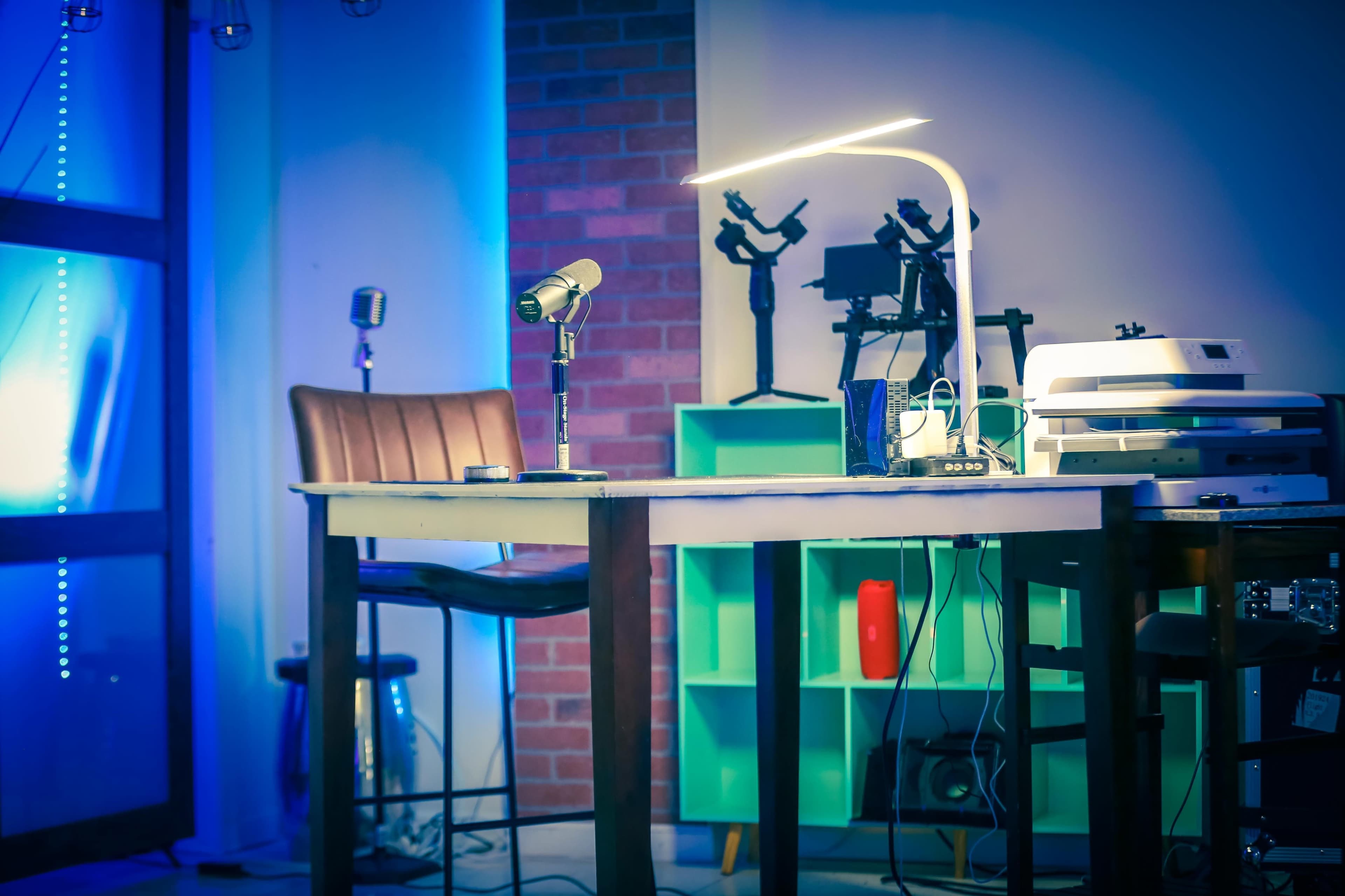 A table with a lamp, a microphone, and various equipment is set against a wall with brick texture and illuminated by blue lighting.