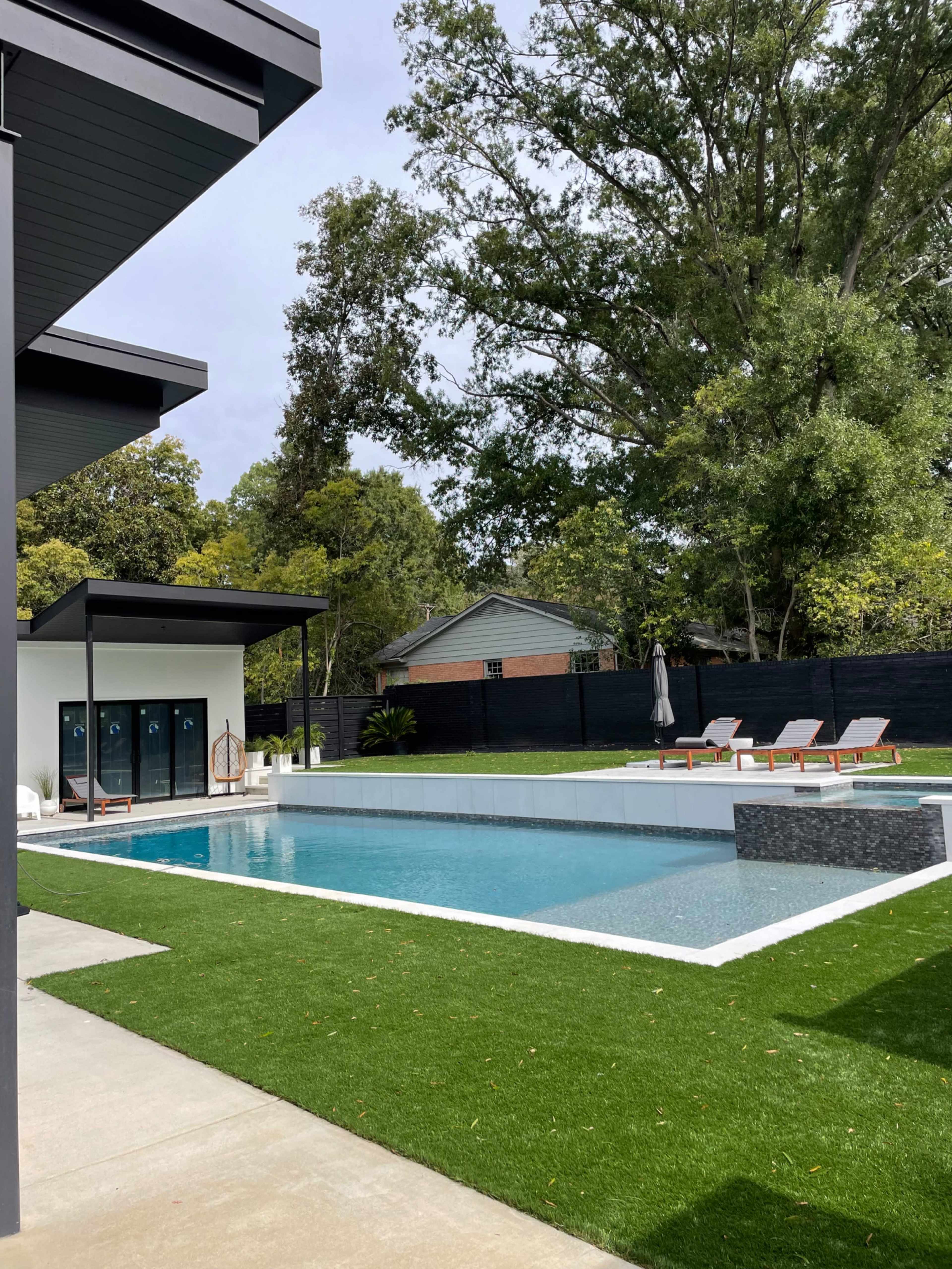 The image shows a modern backyard with a swimming pool, lounge chairs, and a nearby changing area.