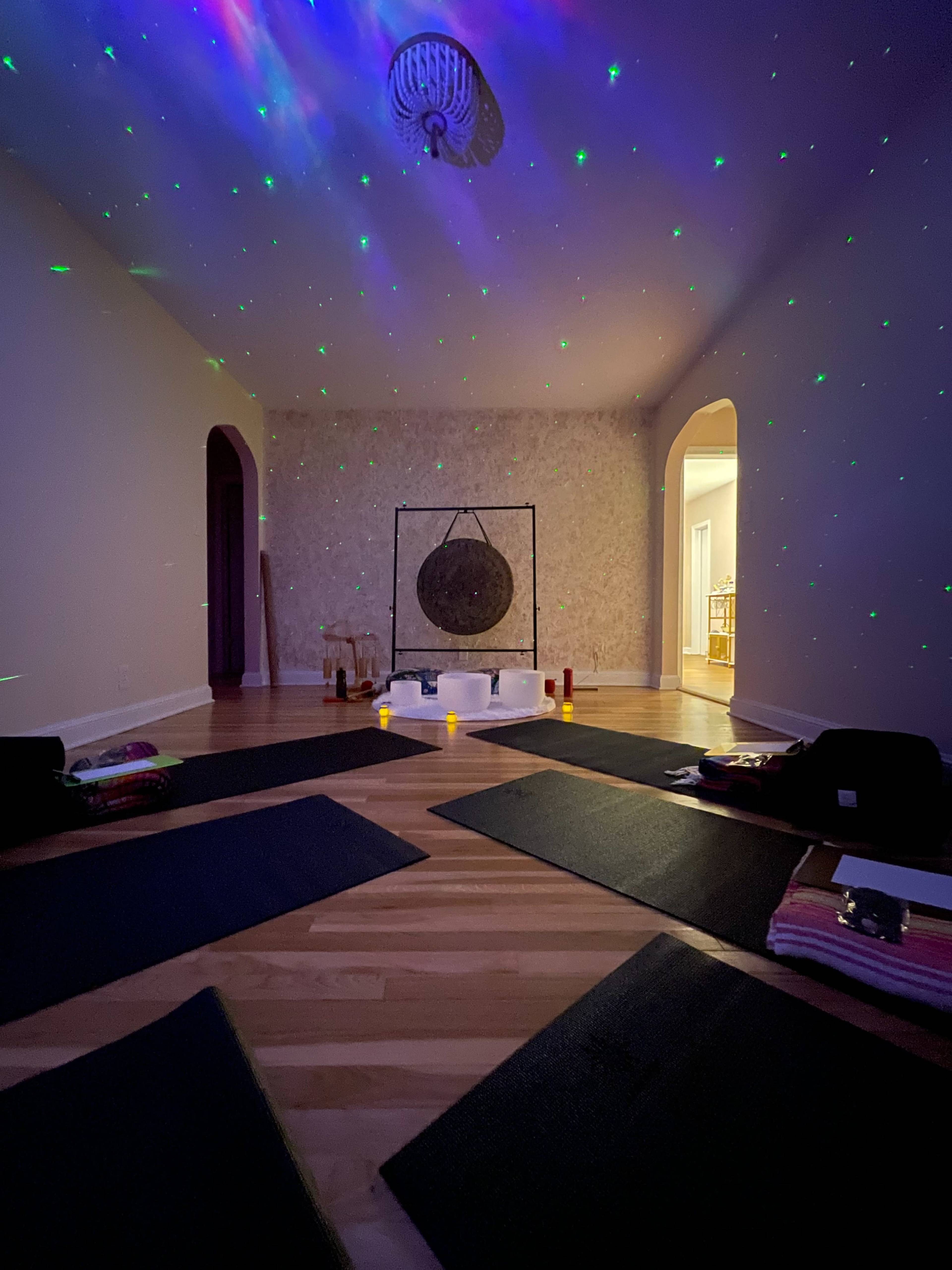 A dimly lit yoga studio features mats arranged on the wooden floor, a gong in the center, and colored lights projecting stars on the ceiling.