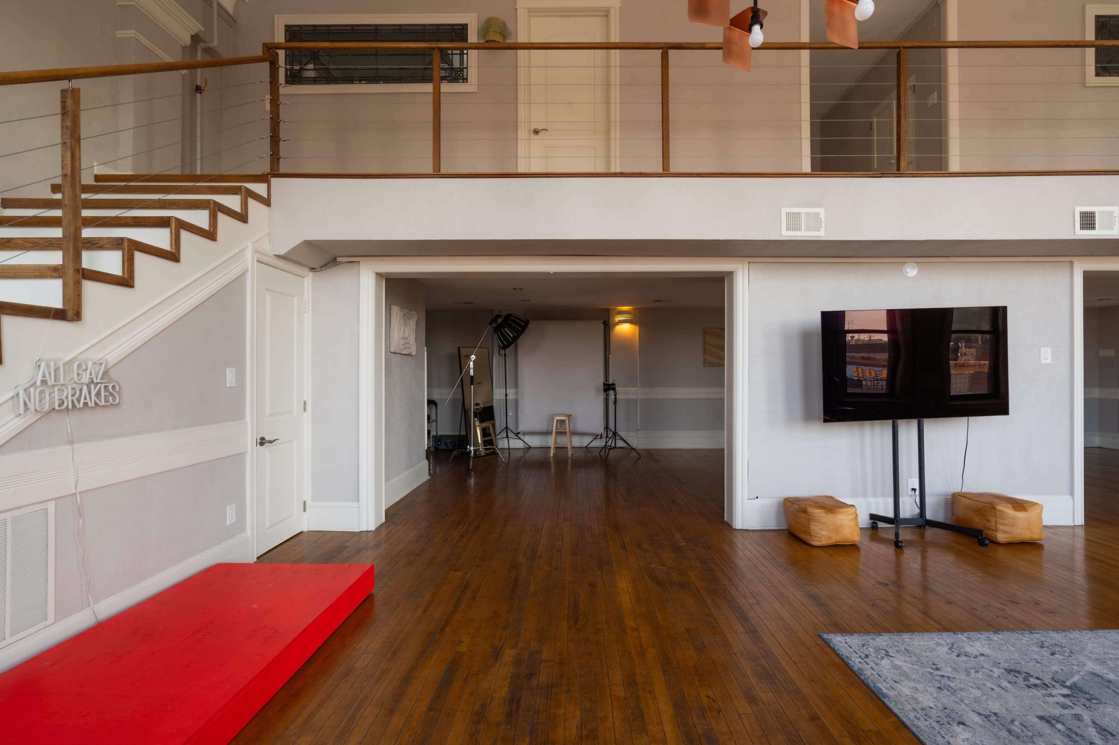 The image shows a spacious, modern living area with a staircase, a red platform, a television on the wall, and a few pieces of furniture.