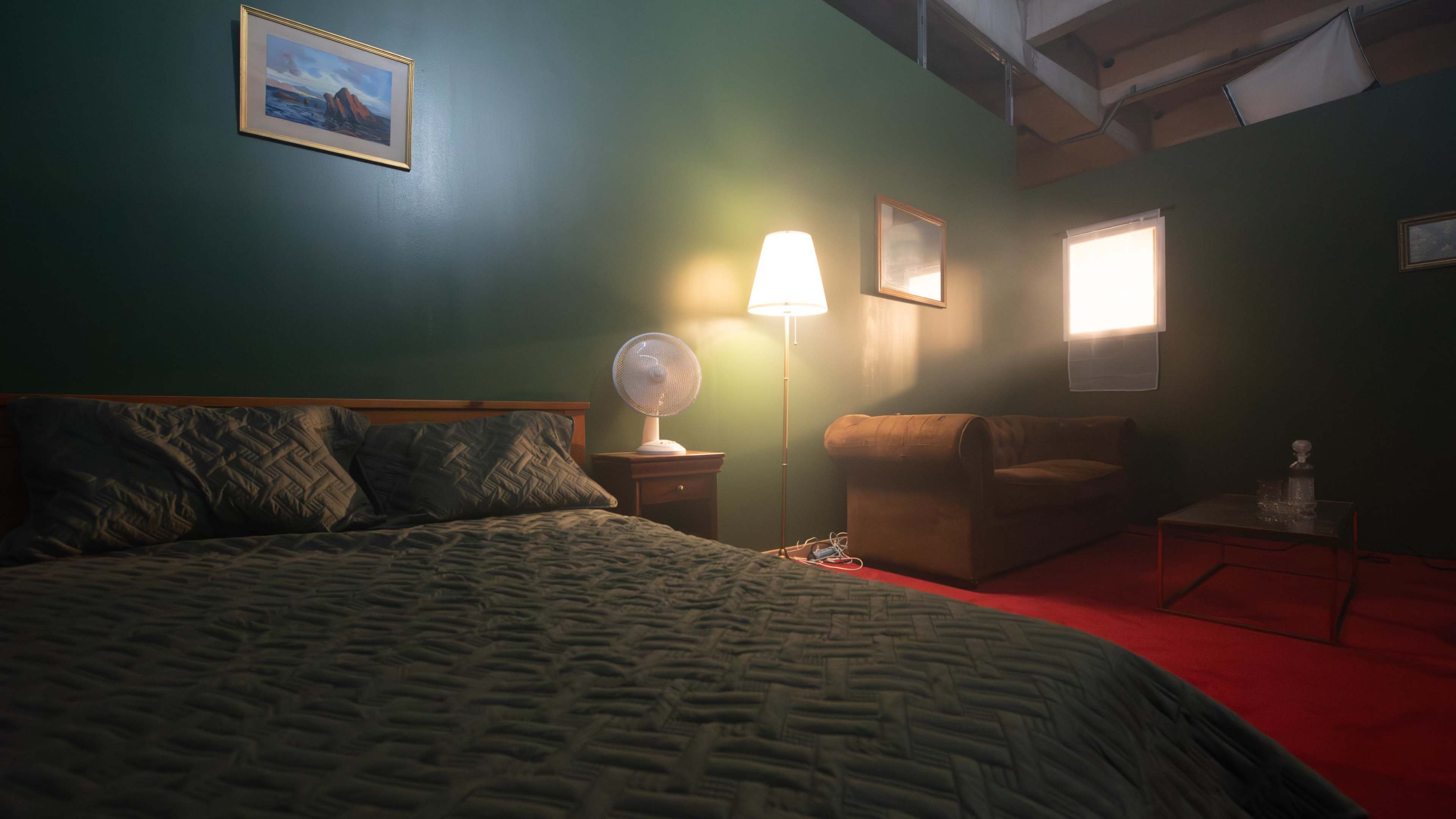 A dimly lit bedroom with green walls, featuring a bed with a patterned cover, a nightstand with a lamp and fan, a sofa, and a coffee table.