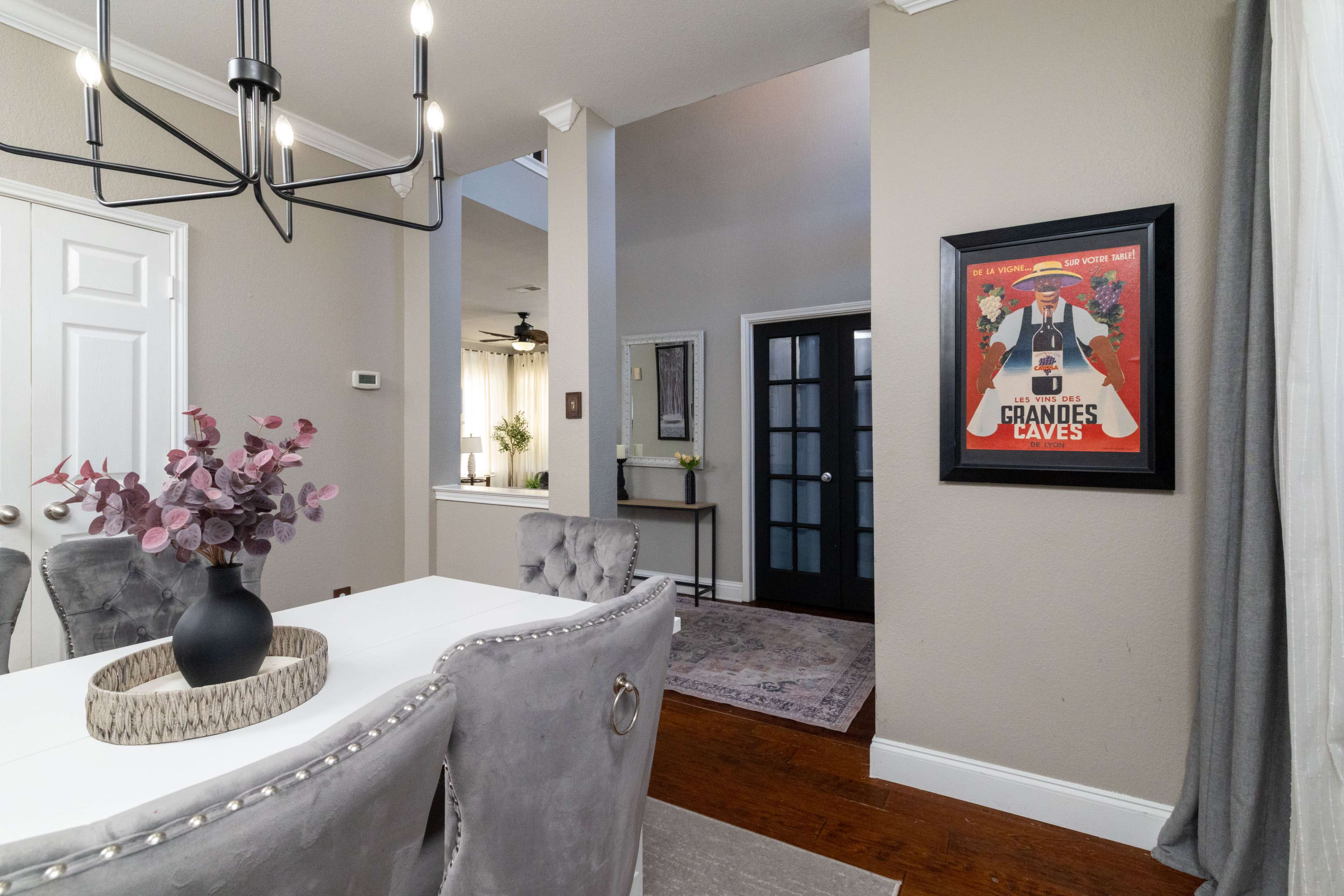The image shows a dining area with a white table, gray upholstered chairs, and a framed poster on the wall, leading to an entrance with a dark door.
