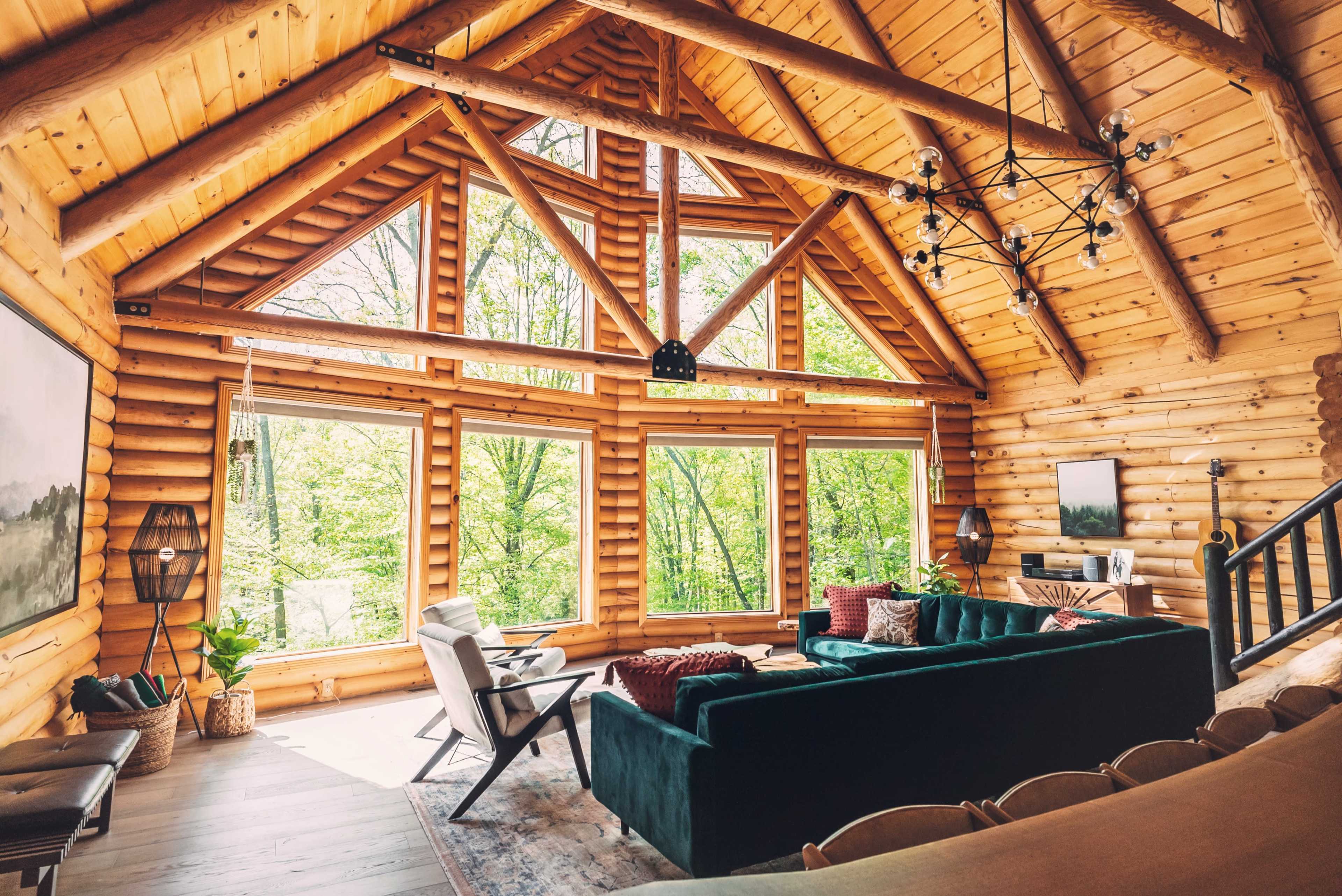 A spacious log cabin interior features large windows, a green sofa, and wooden beams, offering views of a forest.