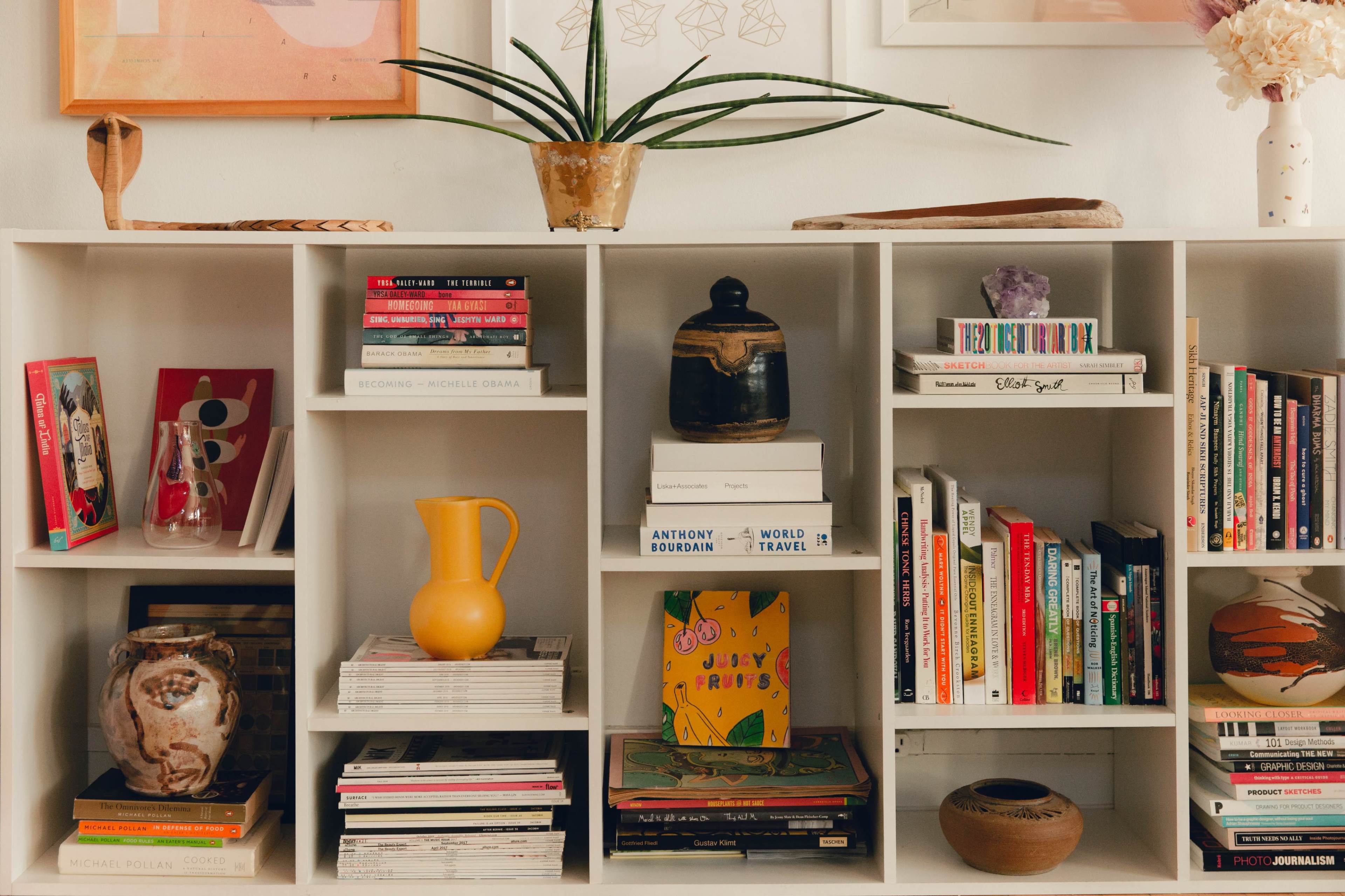 The image features a white bookshelf filled with various books, decorative items, and a potted plant on top.