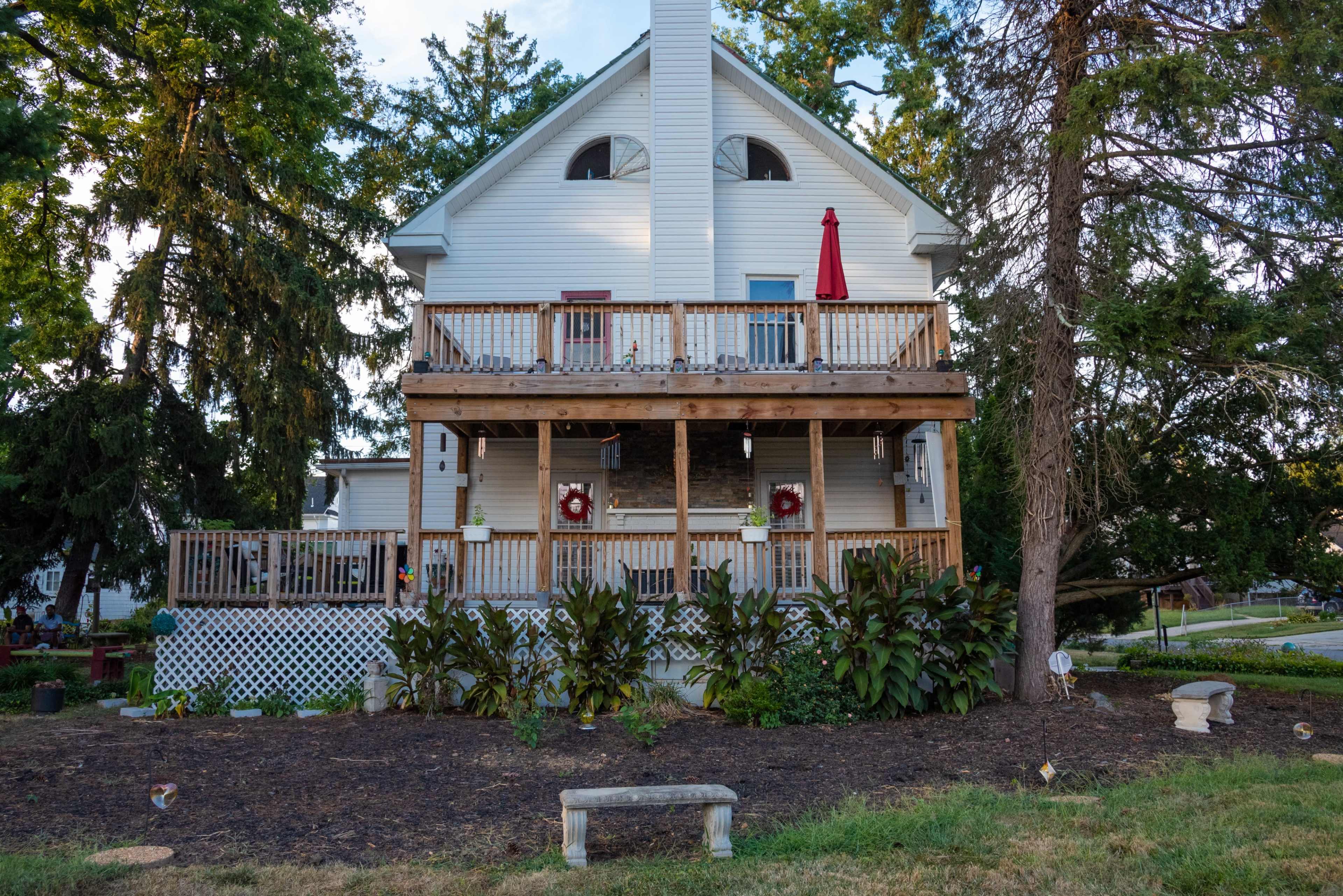 The image shows a two-story wooden house with a large front porch, surrounded by trees and a garden area at the base.